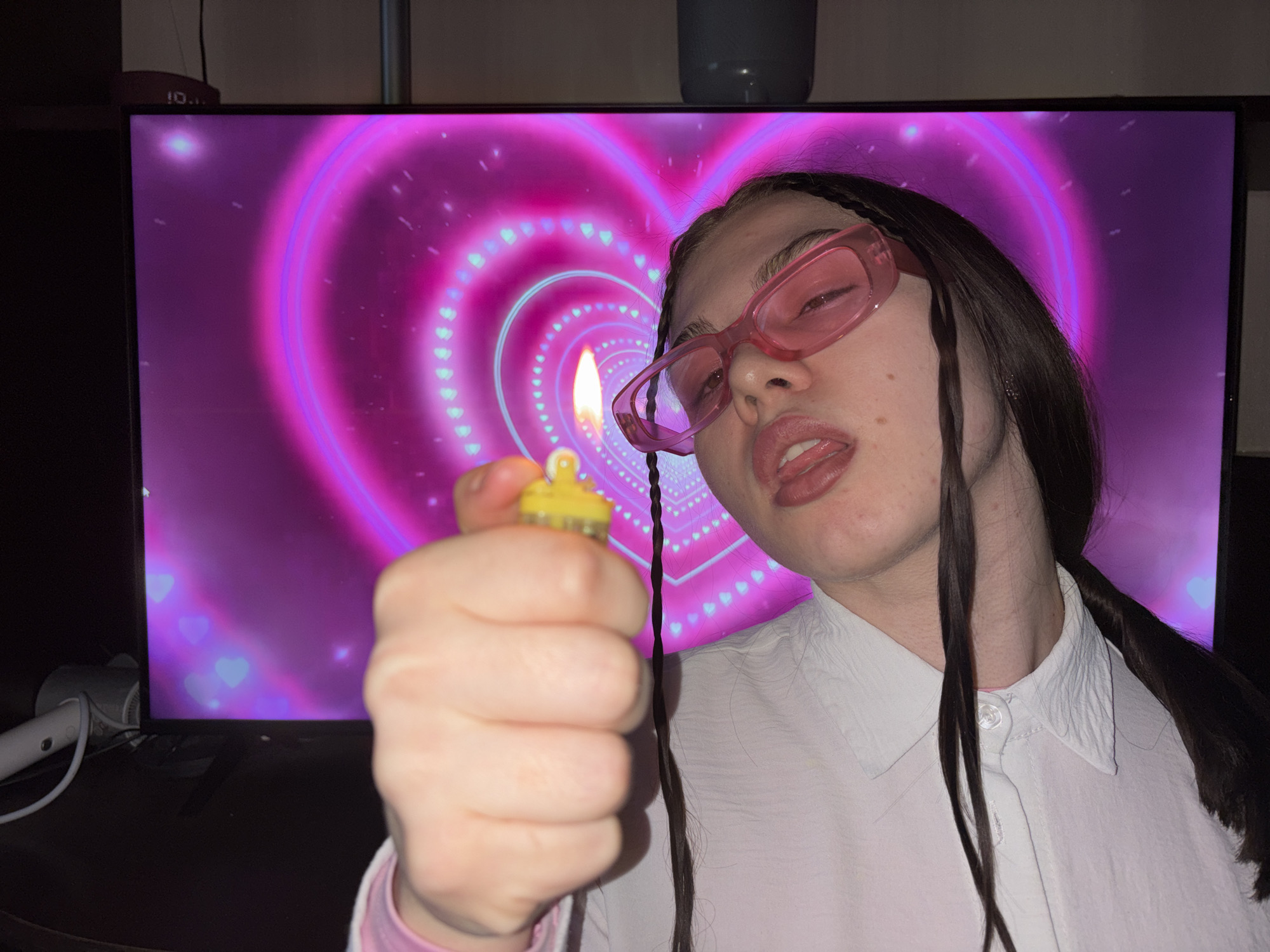 A person is holding a lit lighter in front of a large television screen displaying a pink heart-shaped design. The person is wearing pink glasses and a white shirt. The environment is dimly lit. The time on the device located in the upper-left corner of the television screen is visible as "10:11". The location of Perm, Russia is not discernible from the image.
