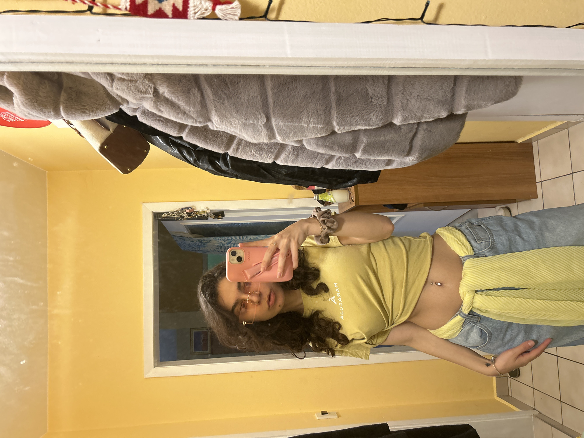 A young woman is taking a selfie in a mirror. She is wearing a yellow t-shirt and blue jeans with a yellow scarf tied around her waist. The woman is holding a pink cell phone. The room has yellow walls, and the floor is tiled. The location appears to be a private residence and not a public place in Sofia, Bulgaria.