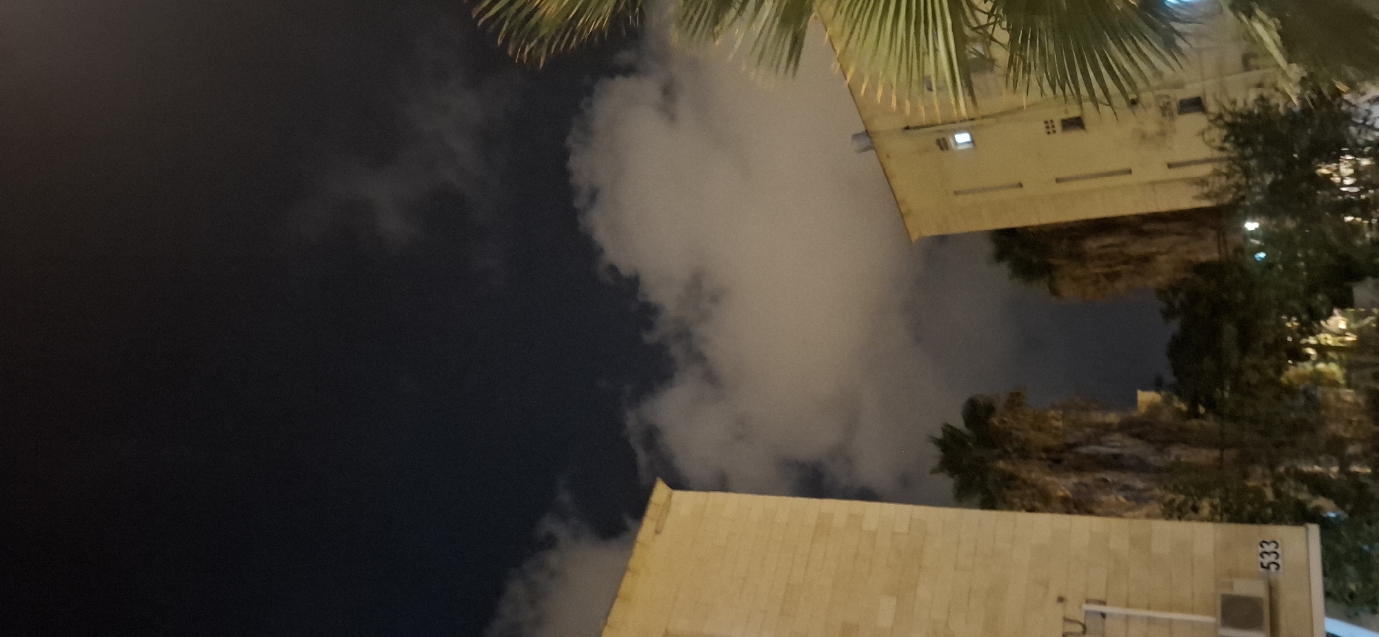 The image captures a nighttime view of a residential building and palm trees in Yerocham, Israel. The sky is dark with scattered clouds illuminated by artificial light. On the left, a portion of a beige-colored building with visible brickwork is shown. A white air conditioning unit and the number "533" are present on its facade. In the center and right of the image, other buildings are visible, with lights on in some windows. Two palm trees stand between the buildings. The foreground features the fronds of a large palm tree on the right side of the frame, partially obscuring the view. The overall scene depicts a quiet urban environment at night.