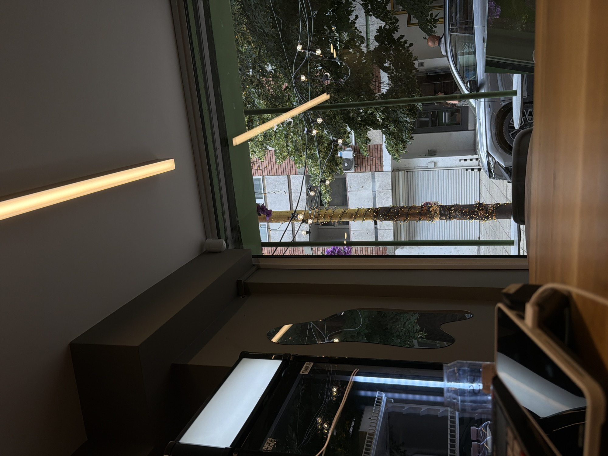 This image depicts an interior scene looking out through a large window onto a city street. The foreground features a wooden table with a black device, possibly a phone or card reader, resting on it. To the left, a glass-fronted display refrigerator with interior lighting is visible. Beyond the window, a street scene unfolds with a pedestrian light pole adorned with numerous small lights. Adjacent to the pole, a modern silver SUV is parked. Trees with green foliage line the street, and in the background, a multi-story brick building with white garage doors is prominent. The environment suggests a commercial establishment such as a cafe or shop on a street in Sofia, Bulgaria.