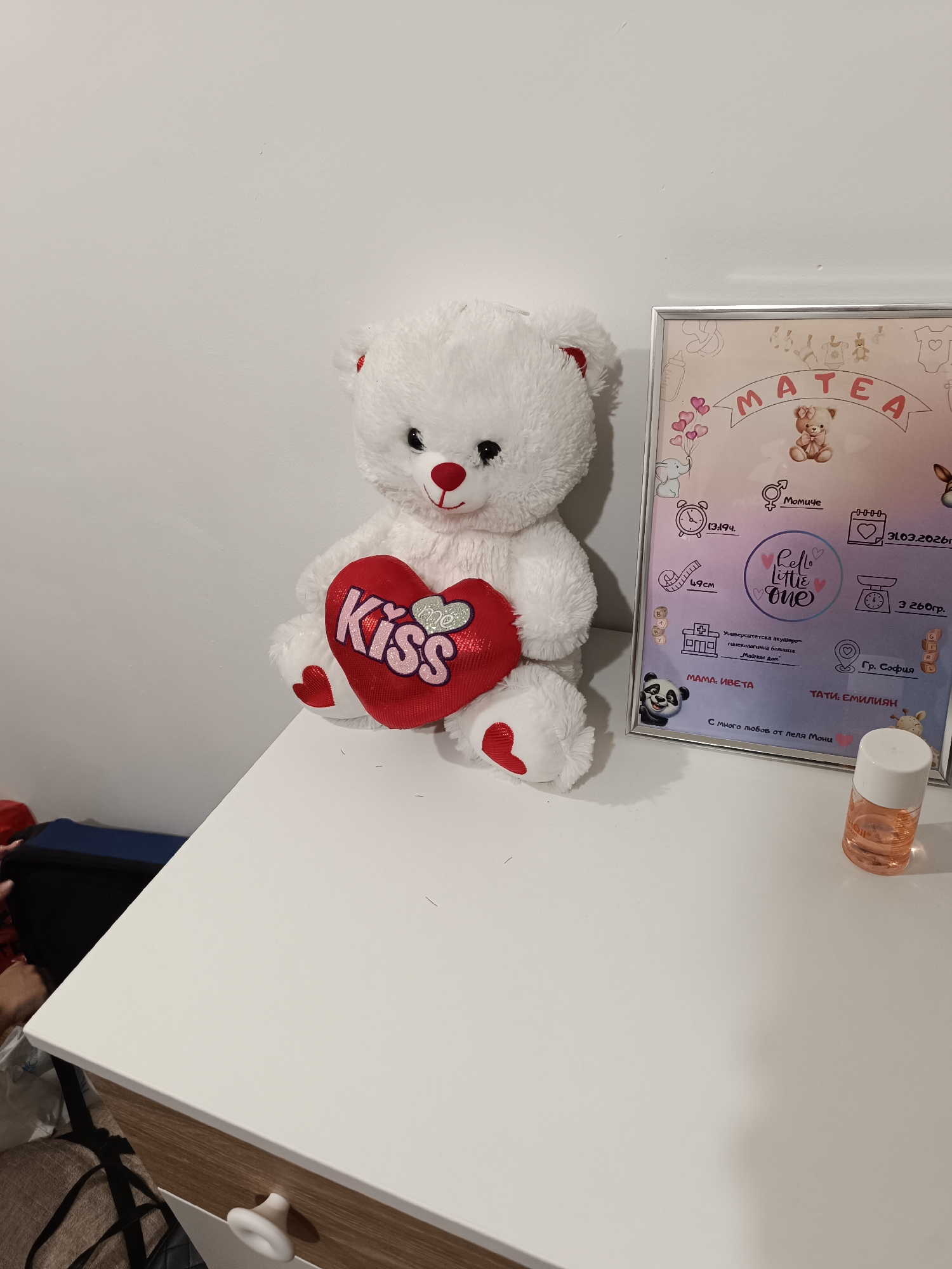 The image captures a scene on a white table. A white teddy bear, holding a red heart-shaped pillow with the word "Kiss" on it, sits next to a framed birth announcement for "MATEA." The announcement includes the date of birth (31.03.2026), birth weight (3260 grams), and a mention of the University Obstetrics and Gynecology Hospital "Maternal Home" in Sofia, Bulgaria, indicating that the birth took place in that city. The announcement also includes the names of the mother, Iveta, and the father, Emiliyan. A bottle of liquid stands on the right side of the table. In the background, a dark blue bag sits on a lower level.