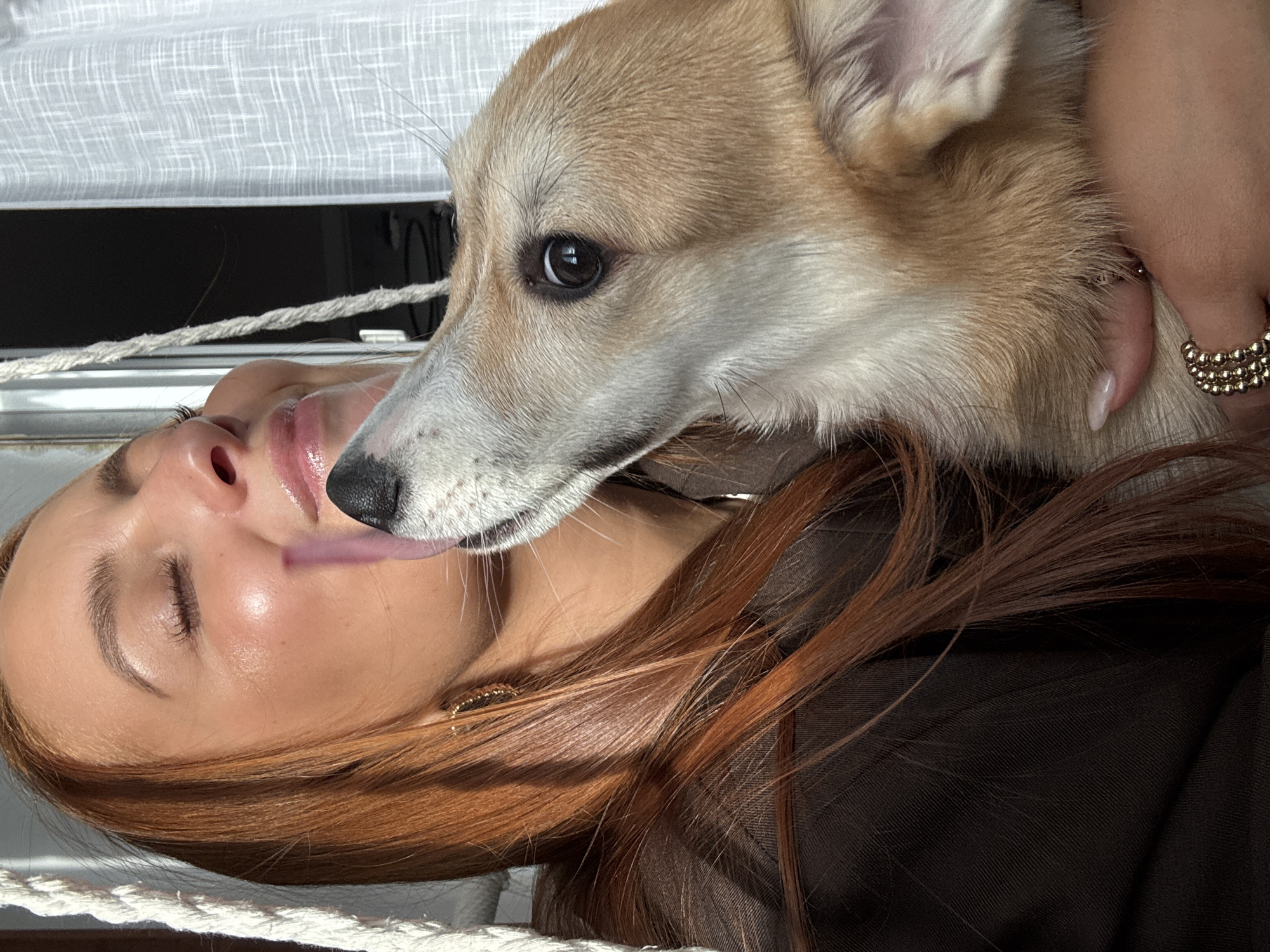 A woman is lying down with a corgi licking her face. The woman has light skin, red hair, closed eyes, and is wearing a brown shirt and gold jewelry. The corgi has tan and white fur and is looking to the left. They are in a well-lit indoor location, likely Chaikovsky, Russia, judging from the tags. A white curtain is in the background.