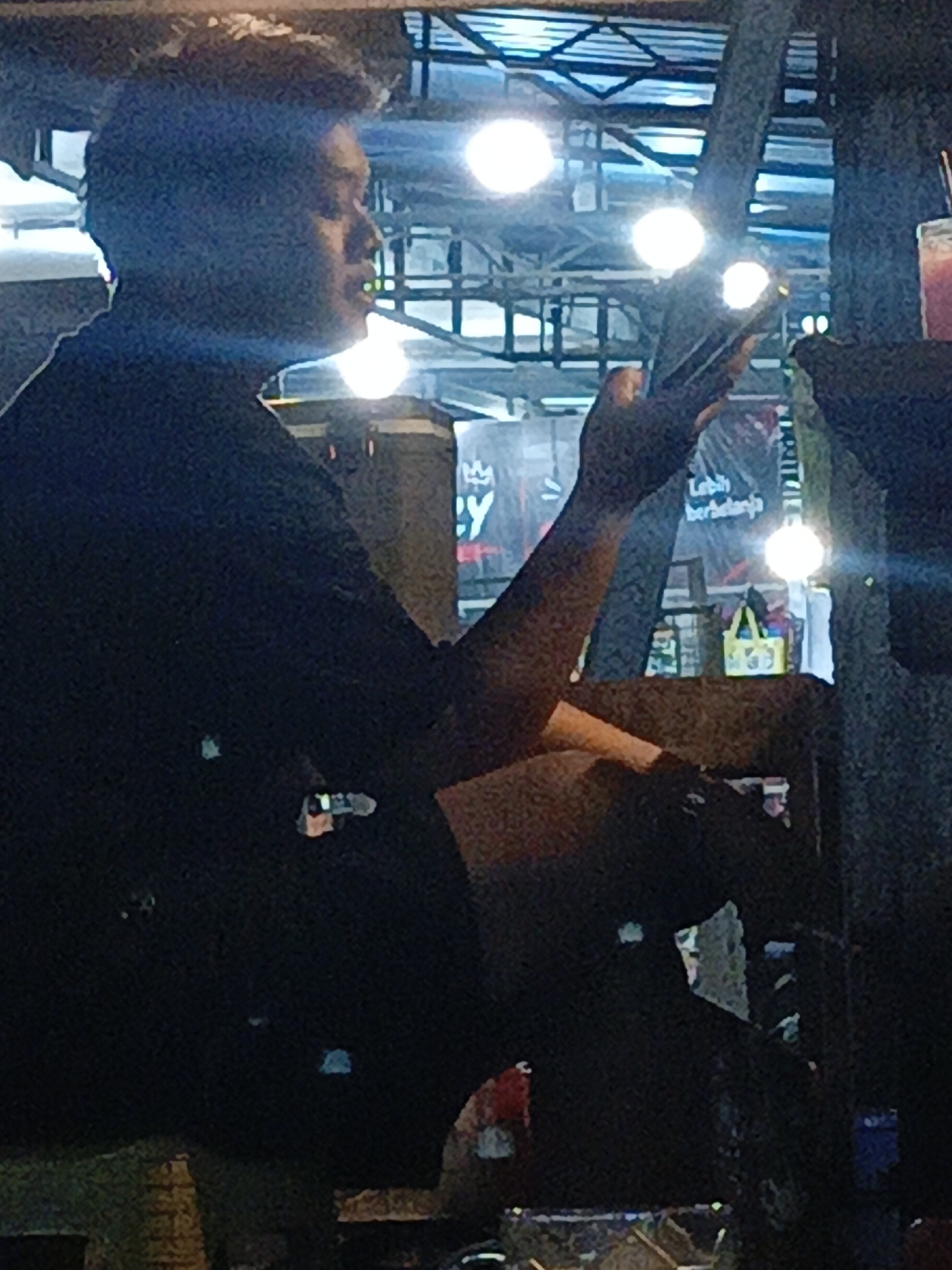 A person in dark clothing is seated, holding a mobile phone in their right hand, angled as if to take a photograph or record a video. The person's face is in profile and partially obscured by shadow, with a slight upward tilt to their head. They are positioned outdoors under a structure with visible lighting fixtures and metal framework overhead. In the background, a banner with text, including "Lebih Berbelanja" (meaning "More Shopping" in Indonesian), is partially visible, suggesting a commercial or market setting. To the right, a bright light source illuminates the scene, and a yellow tote bag with a graphic design is also discernible. The overall lighting is dim, with bright spotlights creating strong contrasts. This scene is located in the city of Purwoharjo, Indonesia.