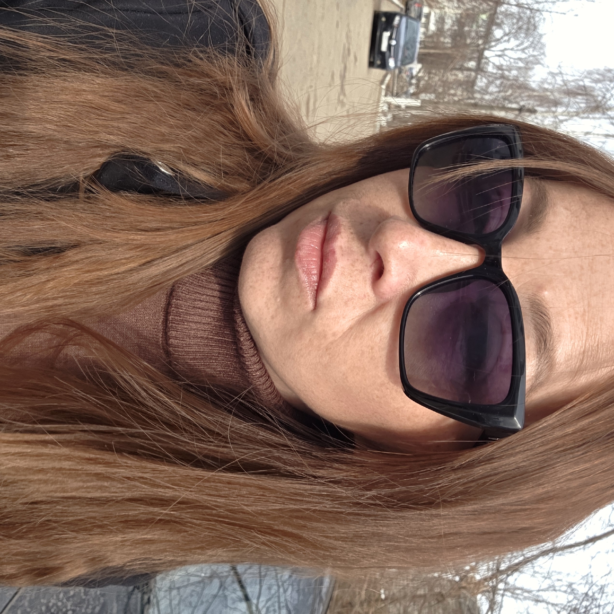A close-up outdoor portrait depicts a female subject with long, reddish-brown hair, wearing dark, cat-eye style sunglasses with purple-tinted lenses, a brown ribbed turtleneck, and a dark outer garment. Her face is turned slightly to the viewer's right, showcasing fair skin with visible freckles on her nose, cheeks, and forehead, and natural pink lips. The background, largely out of focus, reveals an urban street scene in Chaikovsky, Russia, under daylight conditions. A dark-colored sedan is parked on a paved road in the distance to the left, and bare trees are visible, suggesting a season without foliage.