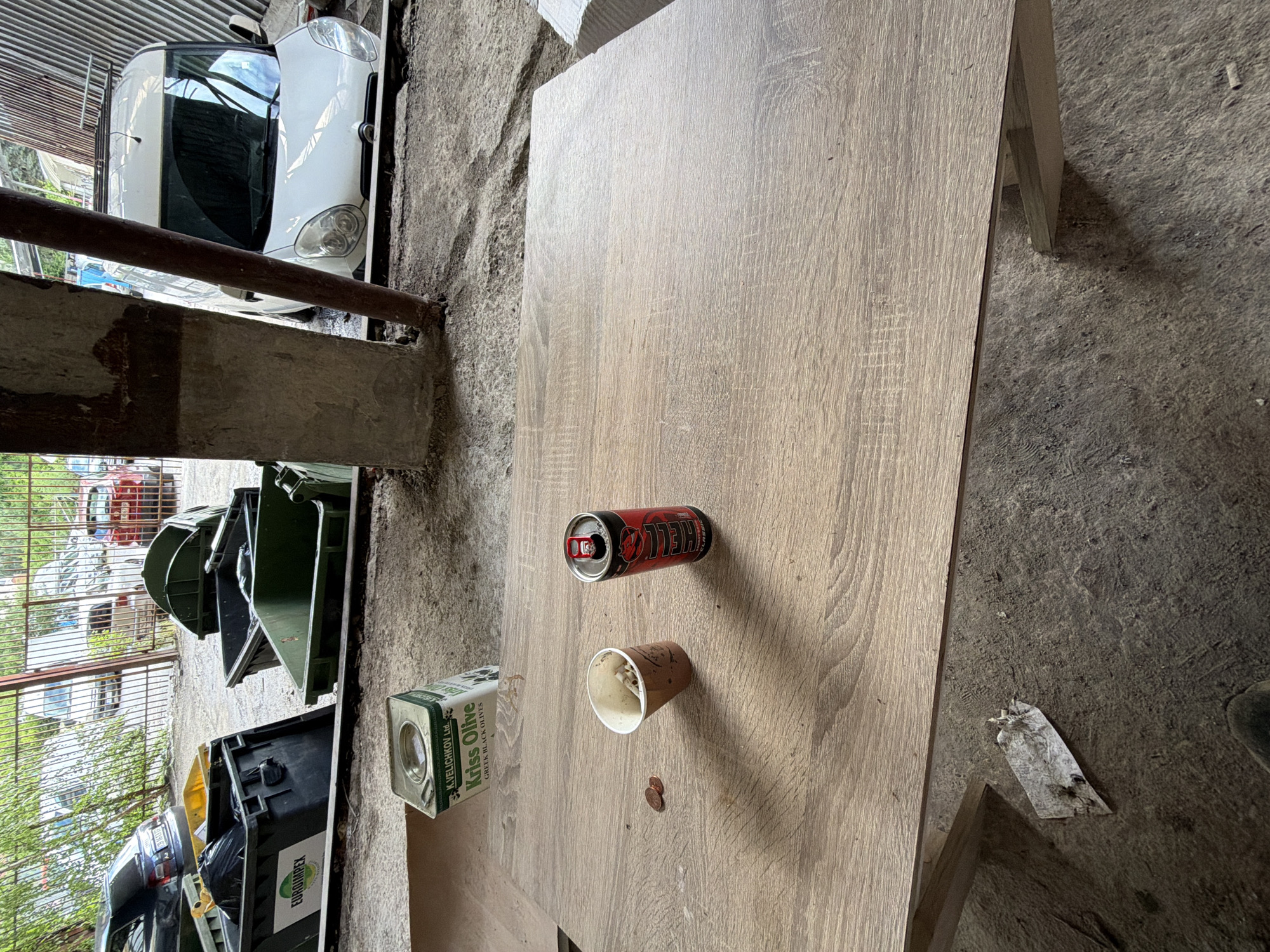 This image, taken in Sofia, Bulgaria, captures a semi-outdoor industrial or storage setting during daylight. The foreground is dominated by a light-colored, wood-textured table, oriented horizontally in the frame.
On the table, several objects are visible: an open red and black "HELL Classic" energy drink can, a disposable paper cup with some residue inside, and two small copper-colored coins. To the left on the table, a rectangular green tin is present, labeled "Kriss Olive GREEN BLACK OLIVES" and additionally bearing the name "K.VELICHKOV Ltd.".
The background reveals a functional, somewhat untidy environment. To the right of the table, the ground is a dirty concrete or asphalt surface. Behind the table, partially obscured by a concrete pillar and a rusty metal bar on the left, a white car, possibly a compact MPV, is visible. Its front end, including headlights and windshield, faces towards the viewer.
Further into the background, beyond the car and a metal fence, several dark green industrial bins are arranged, with the text "EUROMAPEX" clearly visible on one of them. Additional vehicles, including cars of various colors, are parked behind the fence, and some trees and foliage are seen, indicating an open area or yard. The lighting suggests it is daytime, and no people or specific activities are depicted, giving the impression of a static scene in a working or storage facility.