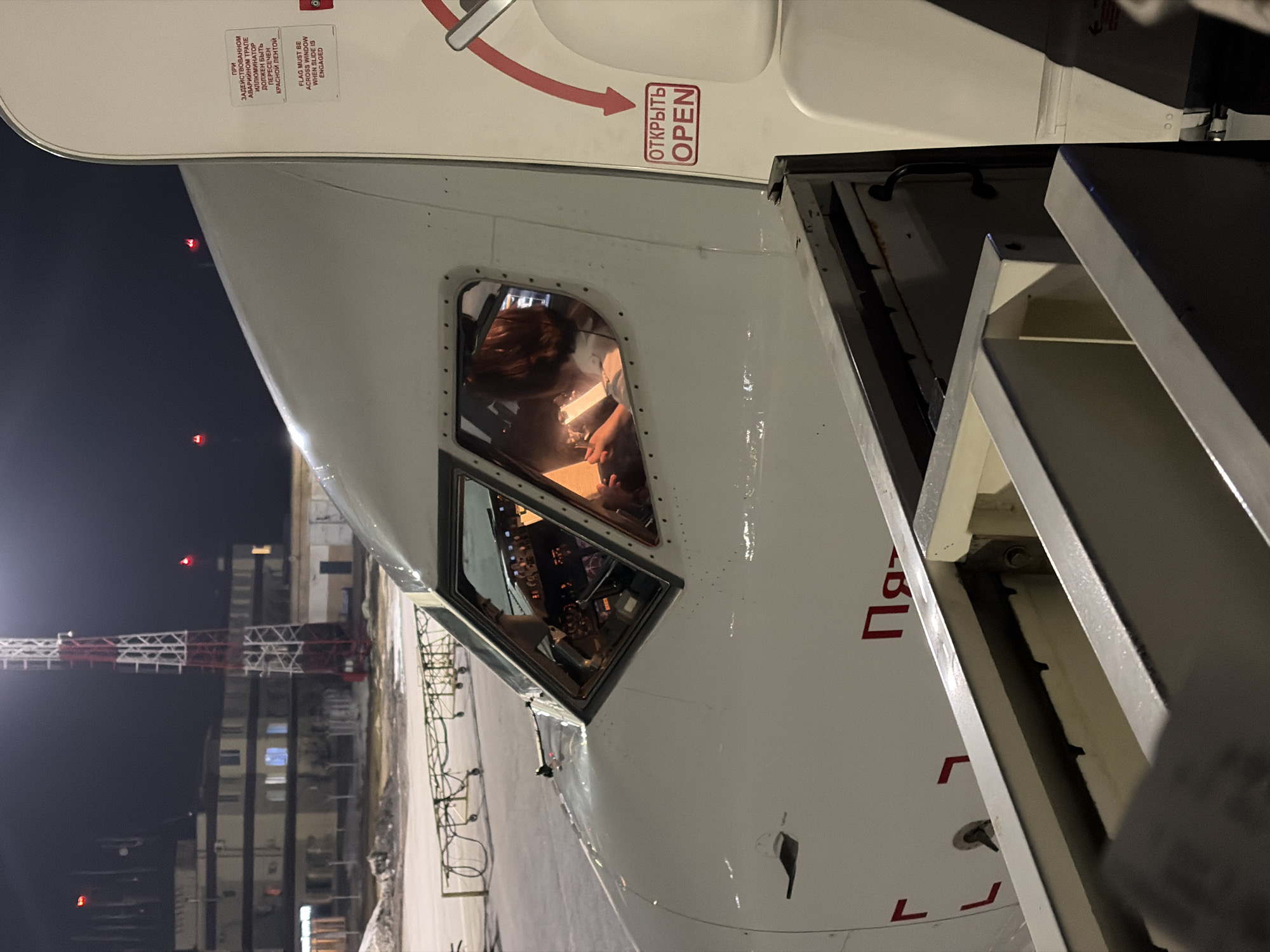 The image displays a nighttime scene on an airport apron in Сокол, Russia, focusing on the front left section of a white passenger aircraft. The aircraft's cockpit is illuminated internally, revealing an individual with reddish-brown hair seated inside, appearing to review a document or interact with an electronic device. The cockpit's instrument panel and controls are discernible. The main entry door on the aircraft's left side is open, exposing its interior mechanism and two visible signs: a red and white sign with "ОТКРЫТЬ OPEN" and a directional arrow, and a white sign above it, in both Russian and English, stating "FLAG MUST BE ACROSS WINDOW WITH SLIDE ENGAGED" related to emergency slide operation. Red text, including the partially visible Russian word "Пять," is present on the aircraft's white fuselage. The airport tarmac exhibits patches of snow or ice. In the background, a multi-story building with illuminated windows stands, adjacent to a tall, red and white lattice communication tower featuring a red warning light at its apex. Additional red lights are visible in the distant dark sky, and yellow wheeled ground service equipment is present on the apron.