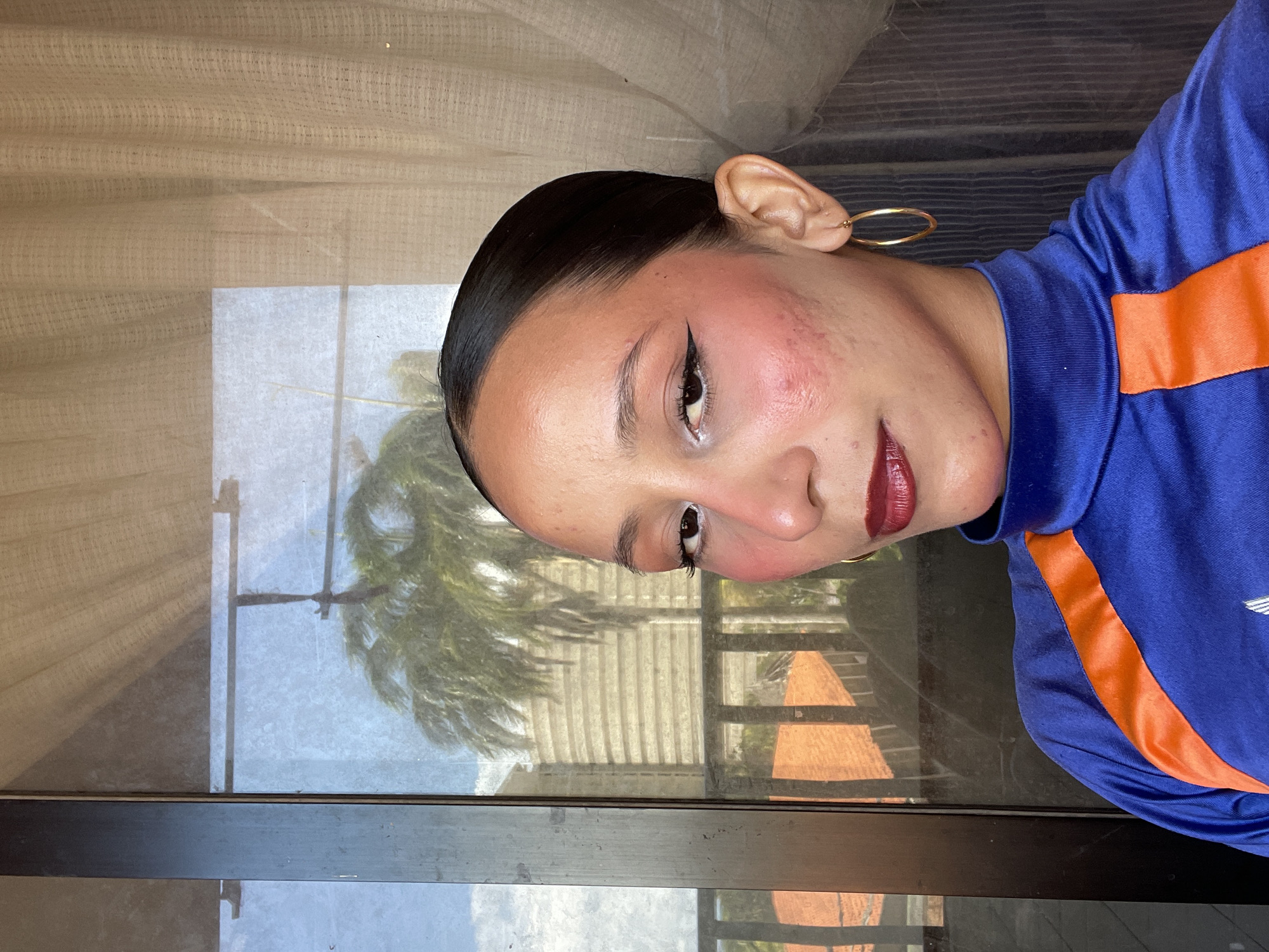 The image shows a close-up of a young woman with dark hair pulled back, wearing a blue and orange athletic top. She has prominent makeup, including winged eyeliner, white eyeshadow, and dark lipstick. There are noticeable blemishes and redness on her cheeks. She is wearing a gold hoop earring. The background features a glass door or window reflecting a palm tree, a building, and a tiled roof, suggesting an urban or semi-urban environment. A sheer curtain is visible on the right. The lighting indicates an indoor setting with natural light coming from outside. This scene could be located in Porlamar, Venezuela, a city known for its tropical climate and coastal architecture, where such views are common.