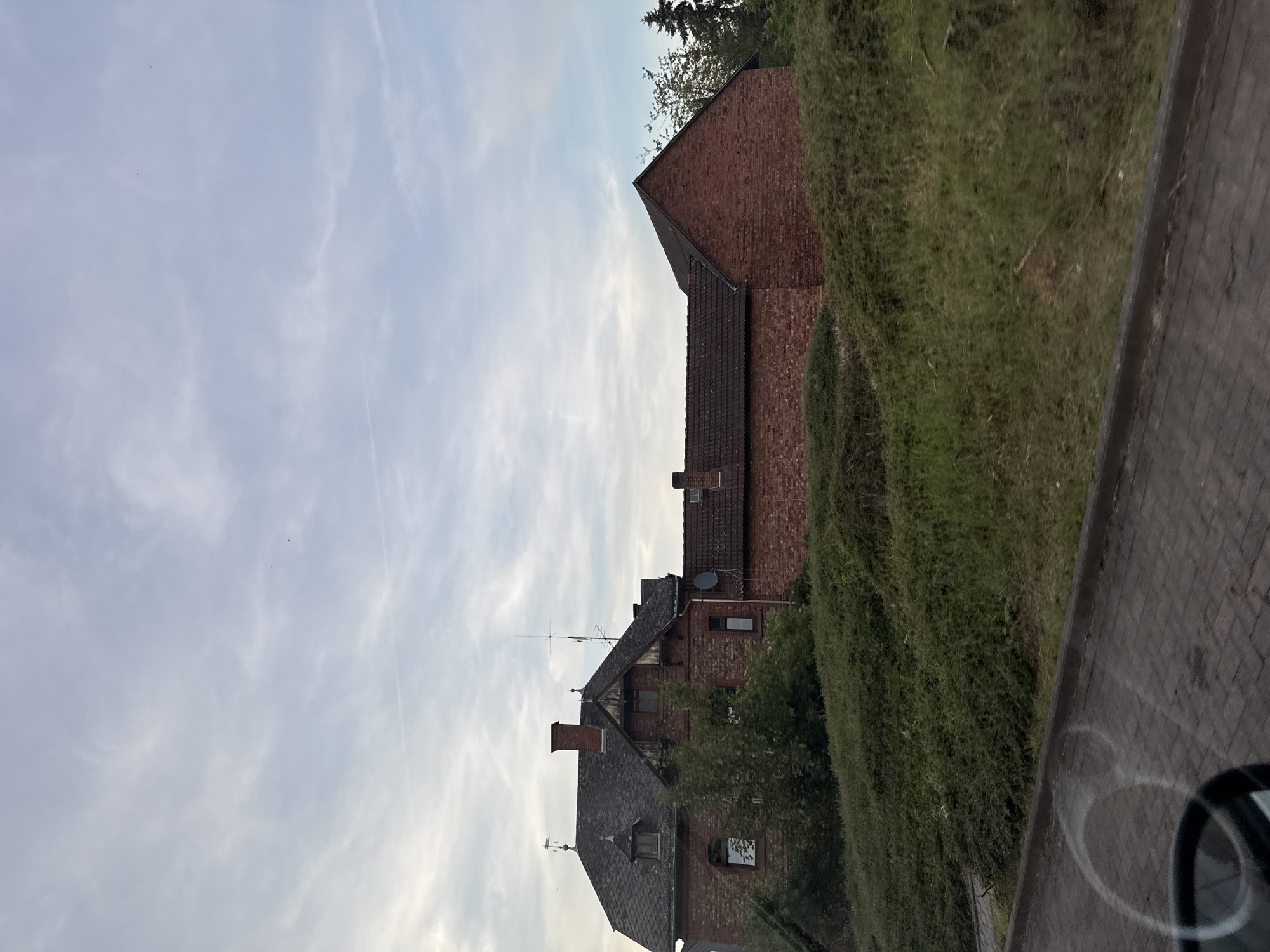 A long, low-lying brick building with a darker tiled roof and a more prominent, older stone and brick section with a steeply pitched roof is situated behind a dense, overgrown hedge. The sky above is overcast with a pale blue hue and scattered clouds. There are some small, dark shapes that appear to be birds in the sky. The foreground shows a paved area and a section of what appears to be a road or sidewalk. The image seems to be taken from a moving vehicle, as indicated by a blurred reflection in the lower left corner. The building has several windows and chimneys, and a satellite dish is visible on the roof of the brick section. Trees are present to the right of the building.