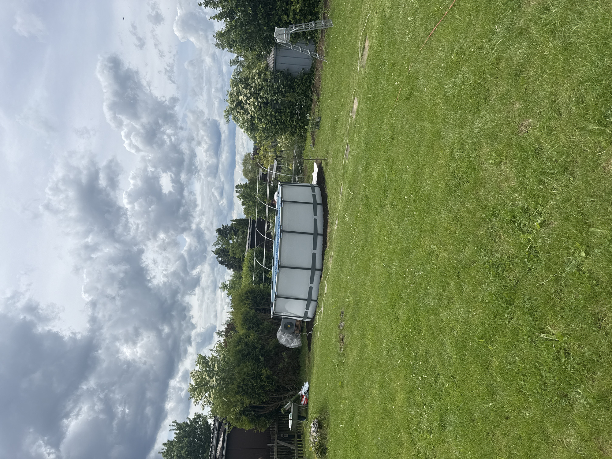 This image displays a residential backyard or garden plot situated in Gotha, Germany. The scene is observed under an overcast sky, dominated by large, grey and white cumulus clouds.
The foreground is comprised of a green lawn with visible grass, weeds, and clover. A light-colored garden hose extends across the lower-mid portion of the frame, running from the left.
In the midground, a large, round, above-ground swimming pool is prominently featured. The pool has a light-colored interior liner and dark grey vertical support beams, and is filled with water. Behind the pool, an incomplete or temporary metallic arched frame structure is visible. To the left of the pool, a green wheelbarrow contains multiple bags and some debris. Adjacent to the wheelbarrow, items covered by clear plastic sheeting rest on a wooden pallet. Further to the right of the pool, a utilitarian shed with grey corrugated metal siding stands. An A-frame metal ladder is positioned upright next to the shed.
The background is characterized by dense vegetation, including various trees and bushes, some displaying white blossoms. On the far left, a dark wooden fence borders the property, with a portion of a building featuring a dark roof visible beyond it. No individuals are present within the scene.