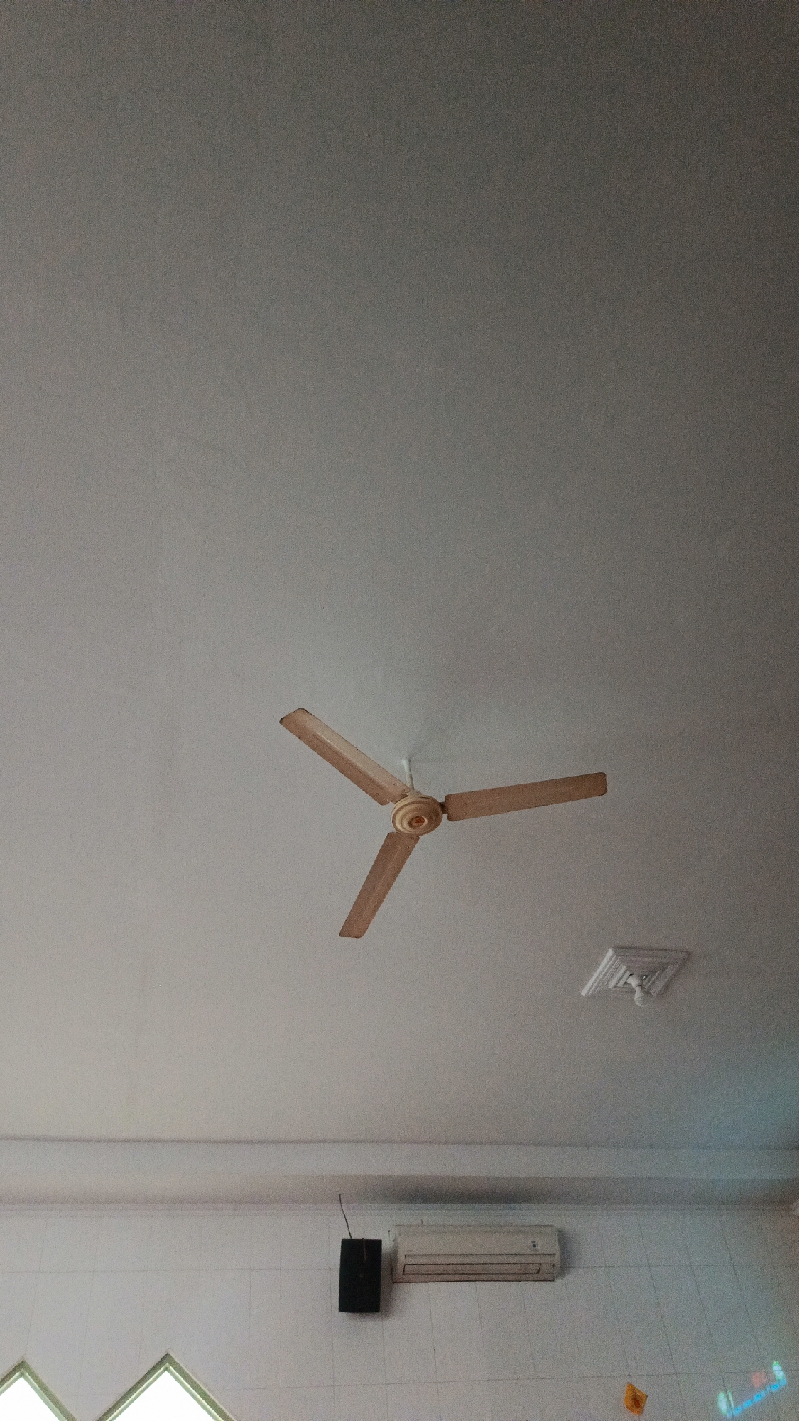 A ceiling fan with three wooden blades is centrally positioned on a white ceiling. To the right of the fan, a square decorative light fixture is attached to the ceiling. Below the ceiling, there is a white tiled wall. On this wall, a black rectangular speaker is mounted, and to its right, a white air conditioning unit is visible. A window with a green frame is partially visible at the bottom left of the image. The overall scene appears to be an indoor space, possibly in a commercial or public building, likely located in a city like Bandung, Indonesia, given the style of the fixtures and the tiled wall. There are no people present in the image.