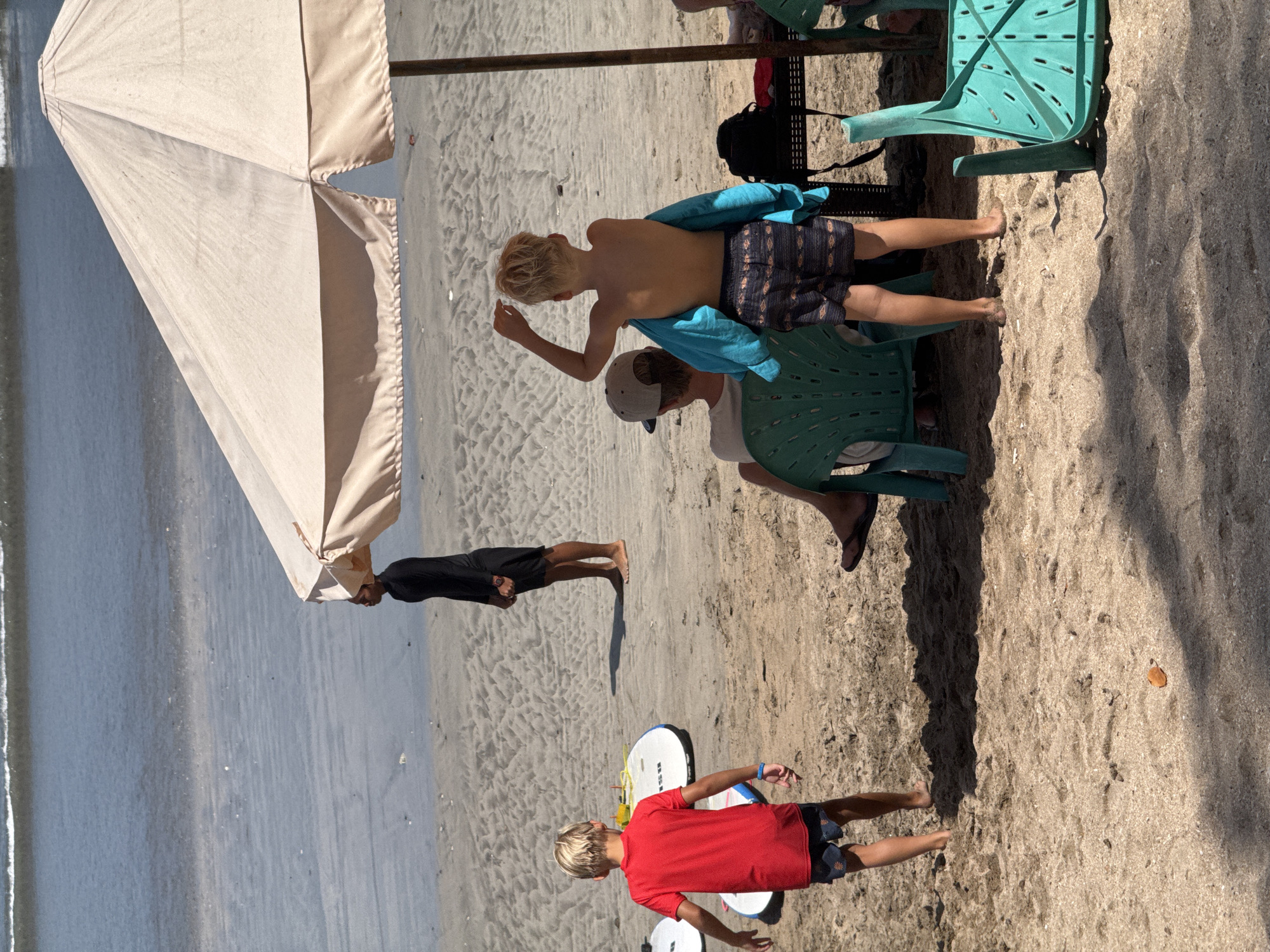 The image depicts a sandy beach scene in Kuta, Indonesia. A person wearing a black long-sleeved shirt and shorts stands near the water's edge, facing away from the camera. In the foreground, two young boys are present. One boy in a red t-shirt and patterned shorts stands on the sand, facing away from the camera. Another boy, shirtless and wearing patterned shorts, stands with a blue towel draped over his shoulders, also facing away from the camera. He has one arm raised. Behind him, a person wearing a baseball cap sits in a teal plastic chair, facing away from the camera. A beige beach umbrella provides shade for some of the area. Surfboards are visible on the sand near the boy in the red t-shirt. The ocean with gentle waves is in the background. The overall environment is a sunny beach with soft sand.