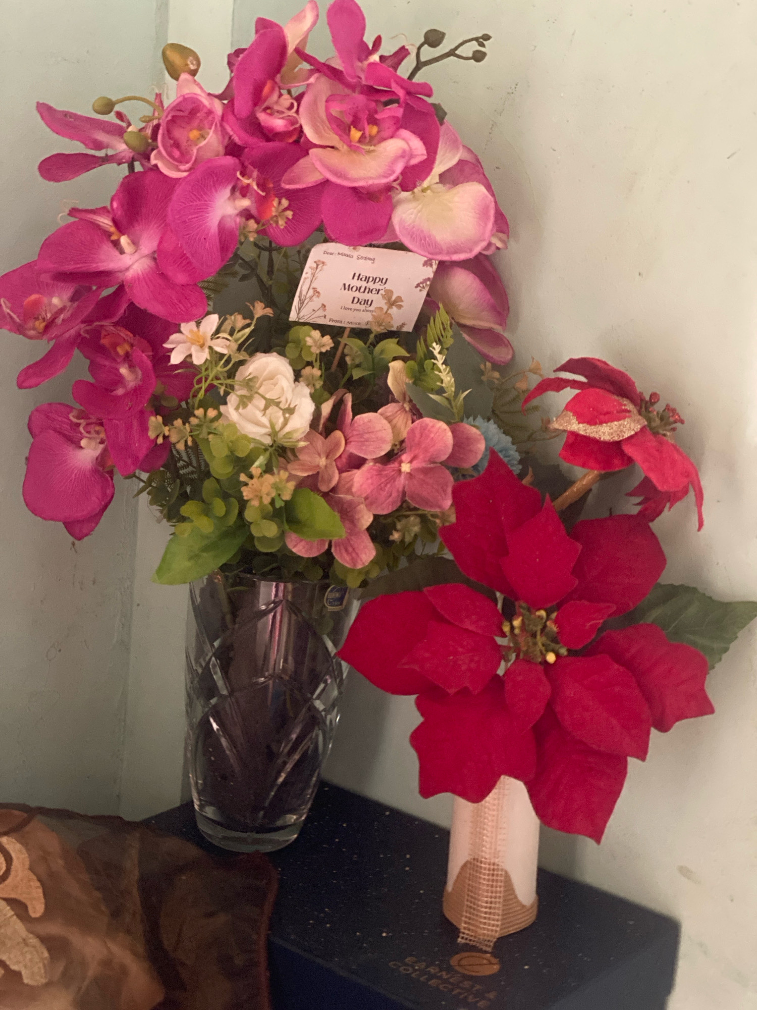 An indoor still life composition features two bouquets of artificial flowers placed on a surface against a light blue wall. The bouquet on the left, predominantly pink orchids and some white and pink filler flowers, is arranged in a tall, clear, faceted glass vase. A small, white card with a floral border is tucked into the bouquet, displaying the handwritten text "Happy Mother's Day I love you always From: Misca". To the right of this, a smaller vase, decorated with a beige burlap ribbon, holds a red poinsettia. The surface beneath the vases is a dark, textured rectangular object, possibly a box, with faint gold lettering that is partially obscured. In the lower left corner, a portion of a dark brown, textured fabric with a subtle pattern is visible. The overall lighting is soft and diffused, creating a slightly hazy atmosphere. The context of Batam, Indonesia, is not discernible from the visual elements of the image.