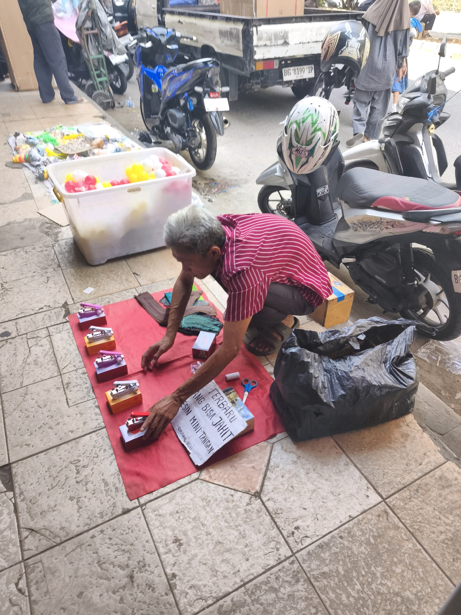 An elderly man with graying hair kneels on a red cloth laid out on a tiled sidewalk in Kelurahan 16 Ilir, Kec. Ilir Timur I, Indonesia. He is wearing a pink and white striped shirt and dark pants. In front of him, on the red cloth, are several small, colorful toy sewing machines, arranged in a row. A piece of paper with Indonesian text, presumably advertising the sewing machines, is also on the cloth, along with a pair of blue scissors and a bundle of fabric. To his right, there is a large black plastic bag overflowing with items. Behind him, a white plastic tub filled with colorful balls sits on the sidewalk, adjacent to a display of other items. In the background, several motorcycles are parked, one of which is a blue scooter. A portion of a white truck is also visible. The overall scene depicts a street vendor selling small items on a sidewalk.