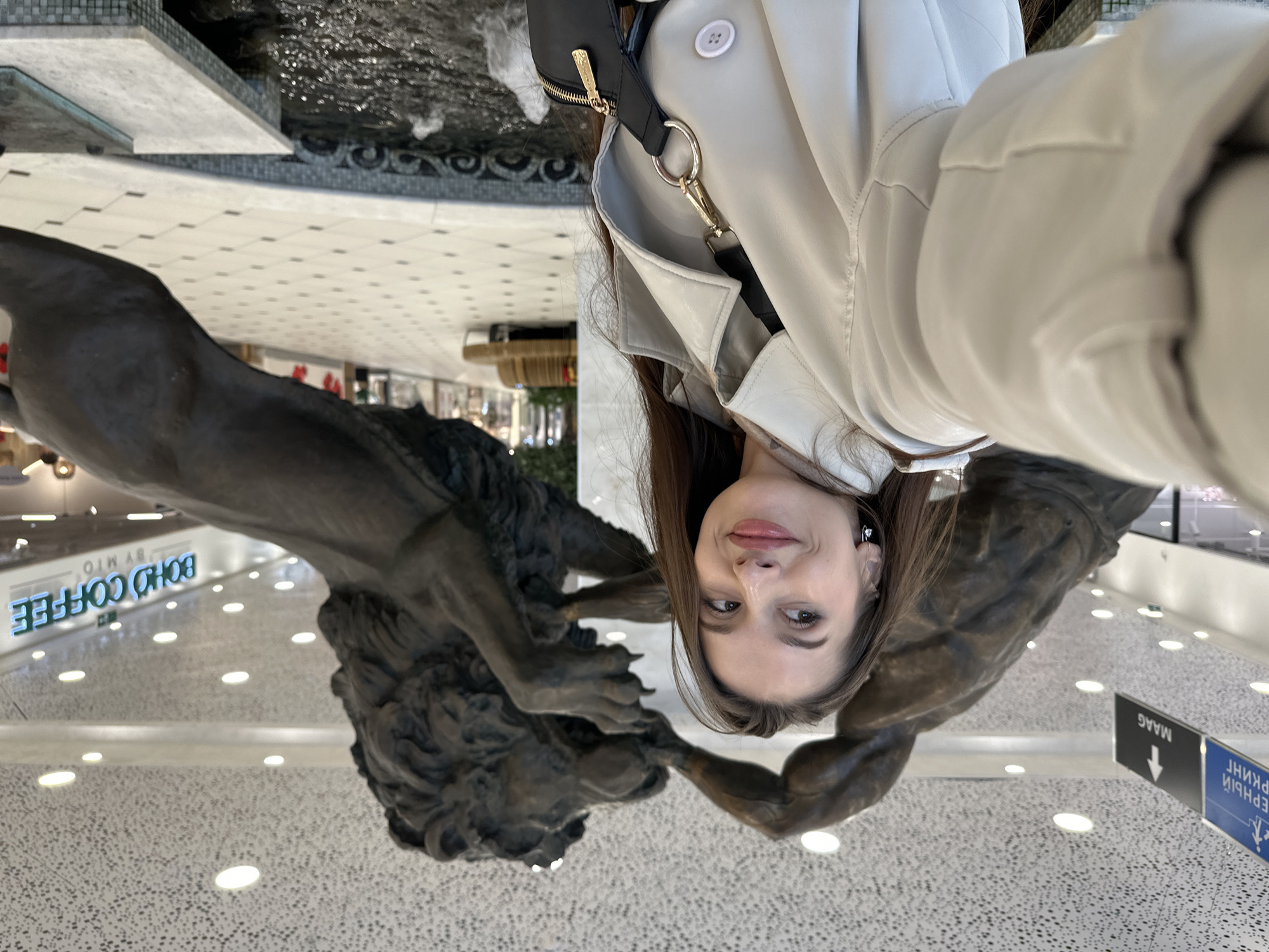 A selfie captures a young woman standing amidst large bronze statues in a spacious indoor setting, likely a shopping mall or public building. The woman, positioned centrally, looks down at the camera with a slight, pleasant smile. She has long, dark brown hair and is wearing a light-colored, possibly cream or beige, trench coat or jacket, with a black shoulder bag featuring gold hardware.
On either side of her and extending into the lower portion of the frame are two impressive dark-colored statues. One appears to be a powerful male figure, possibly engaged in a struggle or interaction with a large lion or lion-like creature, whose head and paws are visible. The statues are highly detailed, depicting strong musculature and texture, suggesting a mythological or heroic theme.
The background reveals a modern, brightly lit interior with a distinctive light-colored, textured floor. To the left, a decorative water feature with dark tiling and splashing water adds to the ambiance. Overhead, numerous recessed lights illuminate the area. Visible in the distance are several signs, some reversed due to reflection. A prominent blue neon sign reads "BOHO COFFEE BY MIO." Another partially visible sign displays Cyrillic text, including "МААГ" (MAAG) and directional information, such as for a "First Floor" (ПЕРВЫЙ ЭТАЖ), consistent with the provided location context of Yekaterinburg, Russia. The scene conveys a sense of a vibrant, contemporary public space.