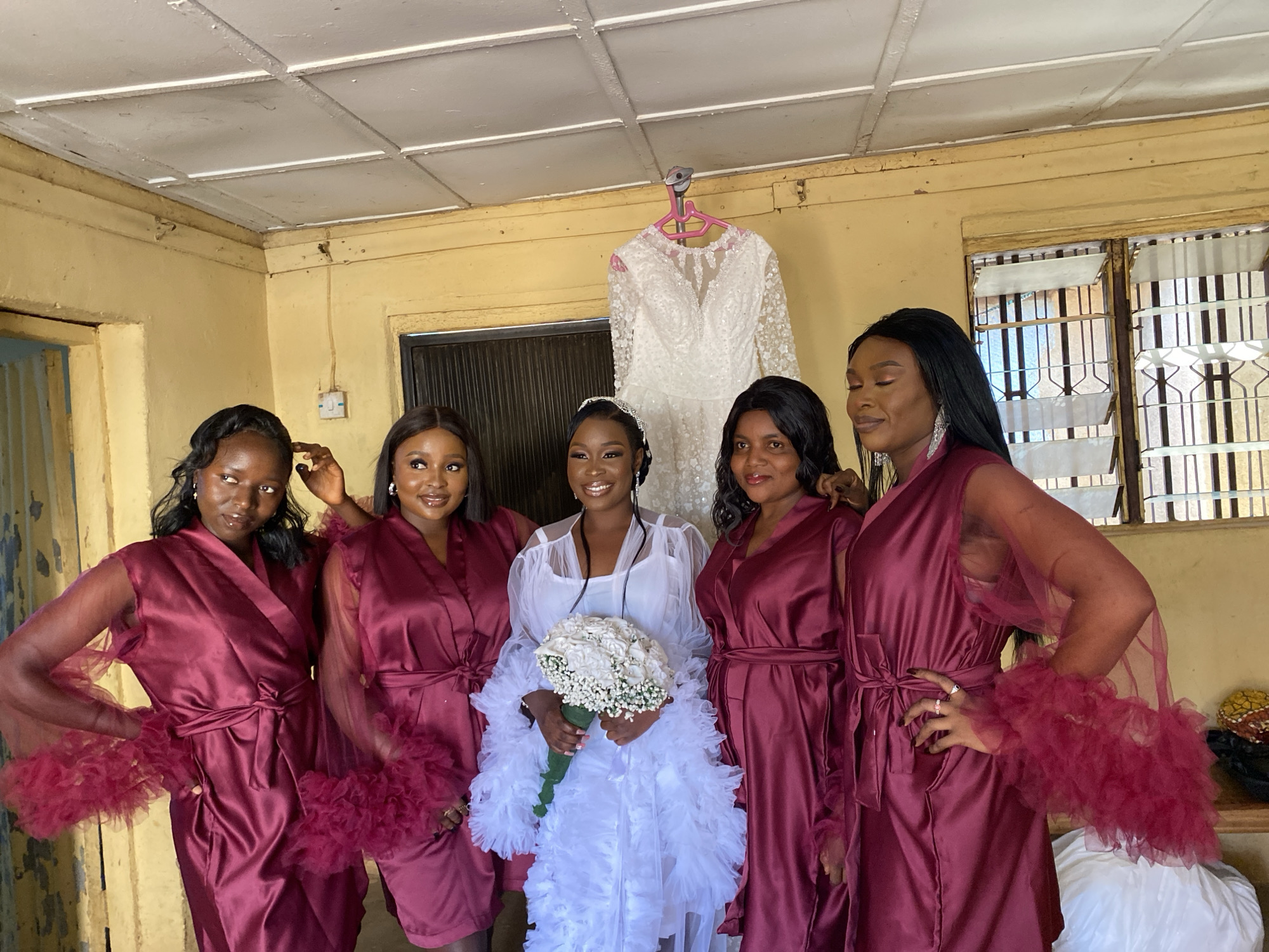 The image captures a wedding party in Vom, Nigeria. Five women are posing indoors. The bride, in the center, wears a white dress with ruffles and holds a bouquet of white flowers. She has a tiara on her head. To her left and right are four bridesmaids, wearing matching burgundy satin robes with tulle detailing on the sleeves. A wedding dress hangs on a pink hanger against the wall. The room has yellow walls and a white ceiling. A window with blinds is visible on the right.