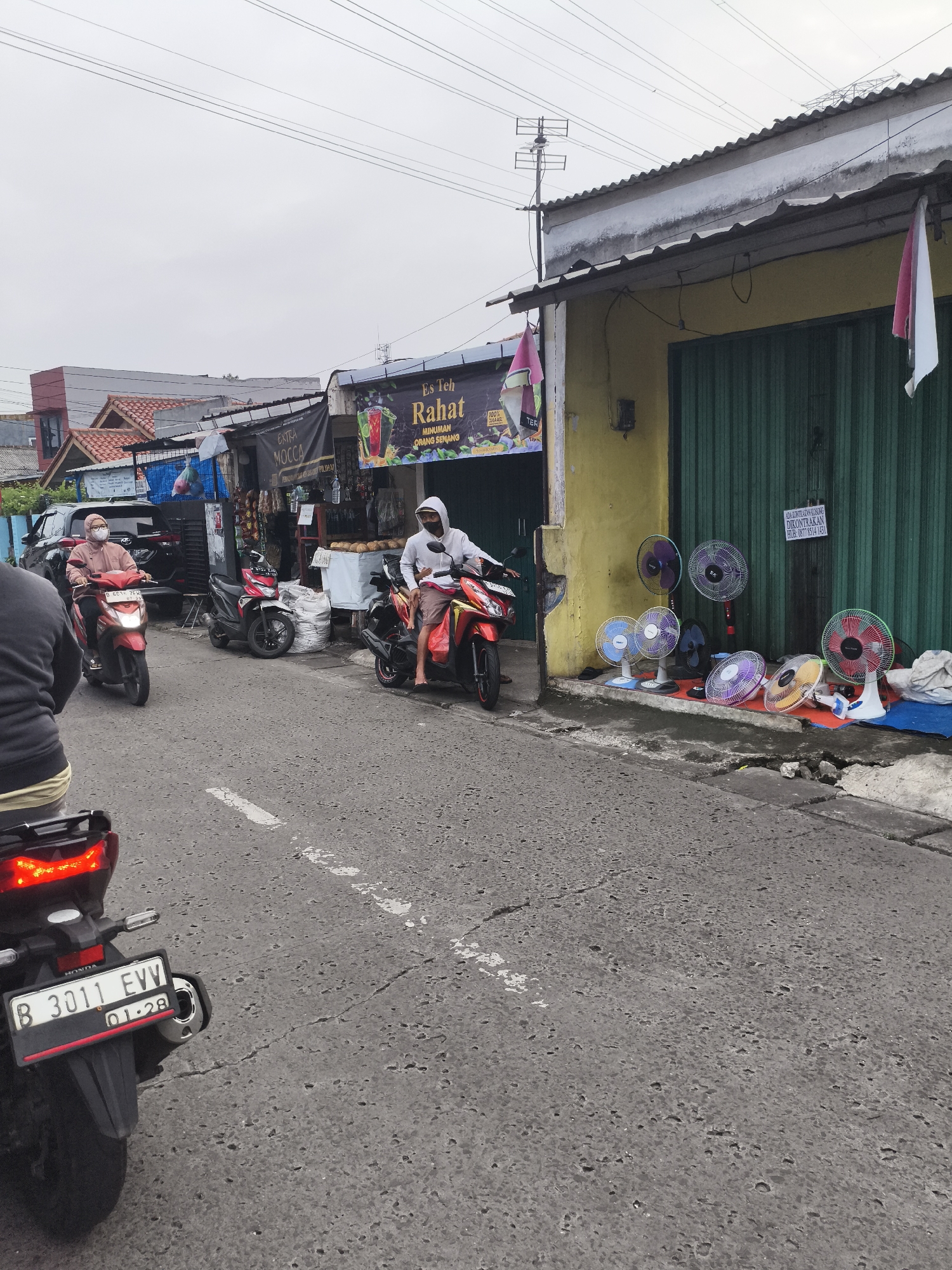The image shows a street scene in Depok, Indonesia. A row of small shops lines the right side of the street. One is selling a variety of electric fans. Another shop sells drinks called "Es Teh Rahat" and "Extra Moccca." A third shop sells various goods. People are riding motorbikes on the street. Indonesian flags hang from one of the shops. The weather appears overcast. The license plate of a motorcycle is visible: "B 3011 EVV" with "01-28" below.