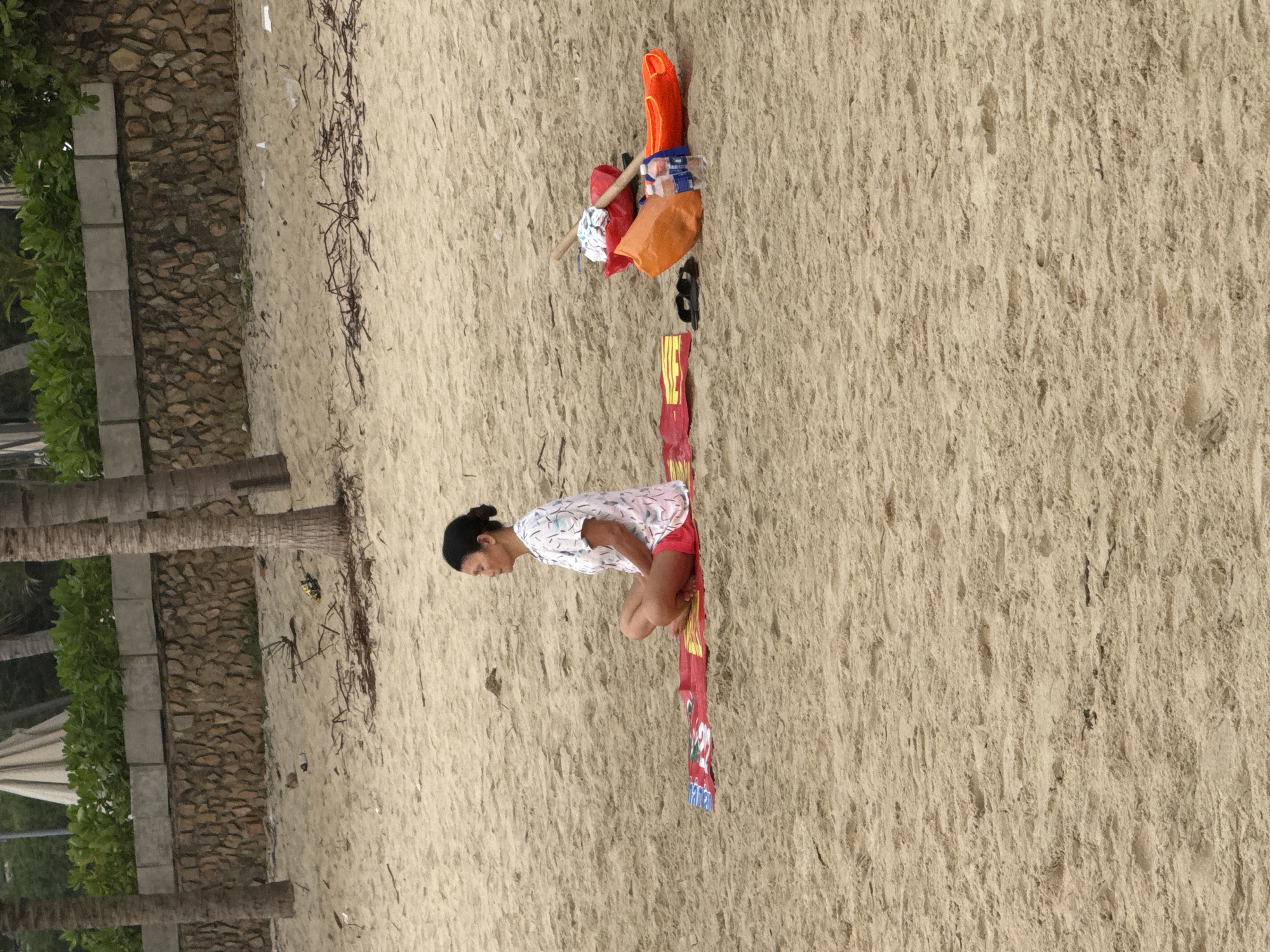 A medium-shot landscape image captures a woman kneeling on a red and yellow mat on a beach. The scene appears to be in Đà Nẵng, Vietnam. The woman has dark hair pulled back in a bun and wears a white patterned shirt and red shorts. Nearby is a collection of orange items, including what appears to be a life preserver, along with other unidentified objects. The beach sand is a light tan color. To the left, a stone retaining wall lines the edge of the beach, with greenery visible behind it, and the trunks of palm trees are also visible. Some dried seaweed is scattered on the sand.