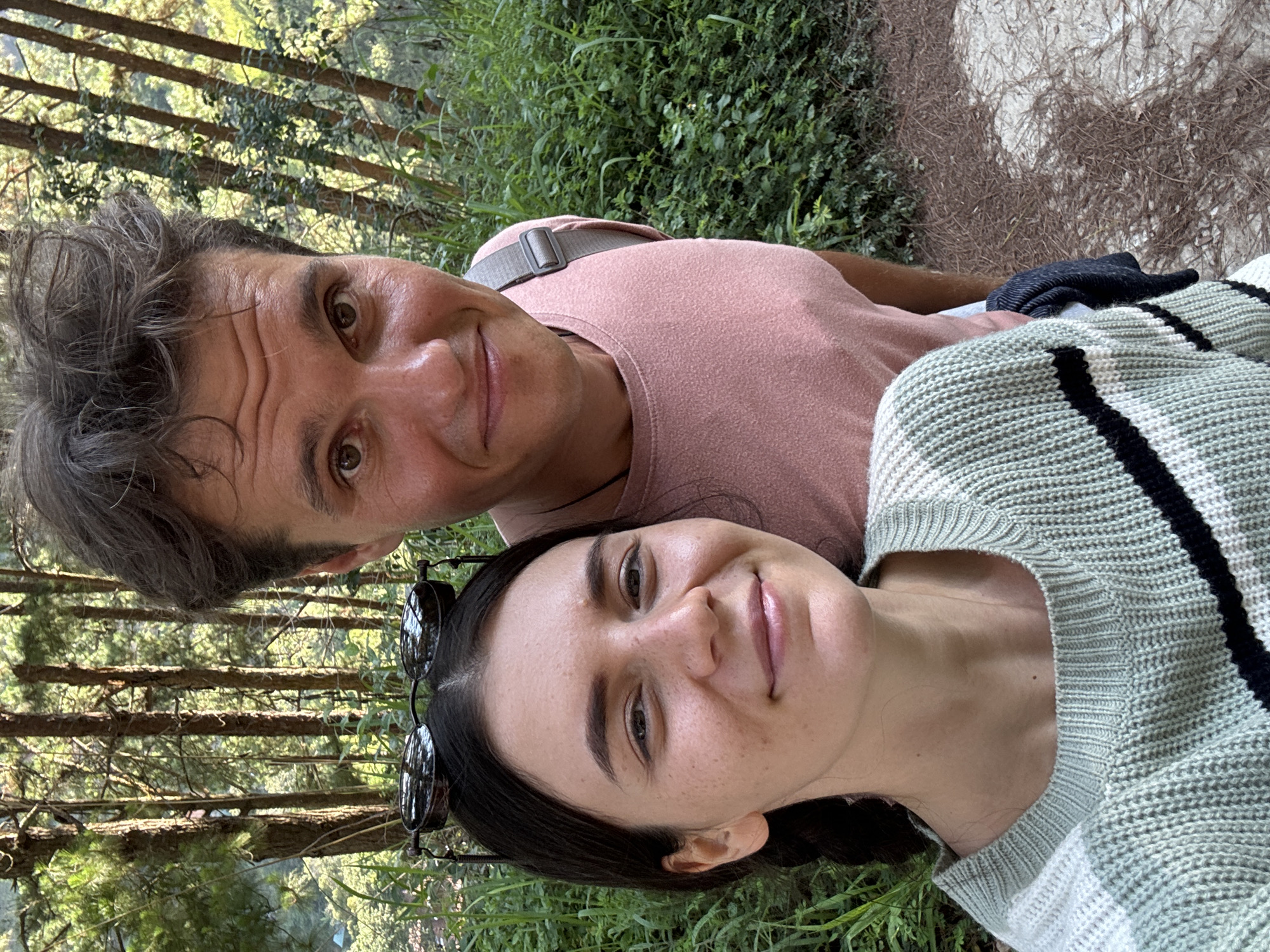 A selfie of a middle-aged man and a younger woman, likely taken in a wooded area in Da Lat, Vietnam.
The man has graying, tousled hair, light skin, and brown eyes. He is wearing a pink t-shirt and a gray strap is visible across his chest. He is positioned above the woman.
The woman has dark, braided hair, fair skin, and is wearing sunglasses. She is wearing a light green sweater with black stripes.
The background includes a dense forest with tall trees and green foliage. Part of a concrete area is visible at the top right.
The subjects appear to be lying on the ground.