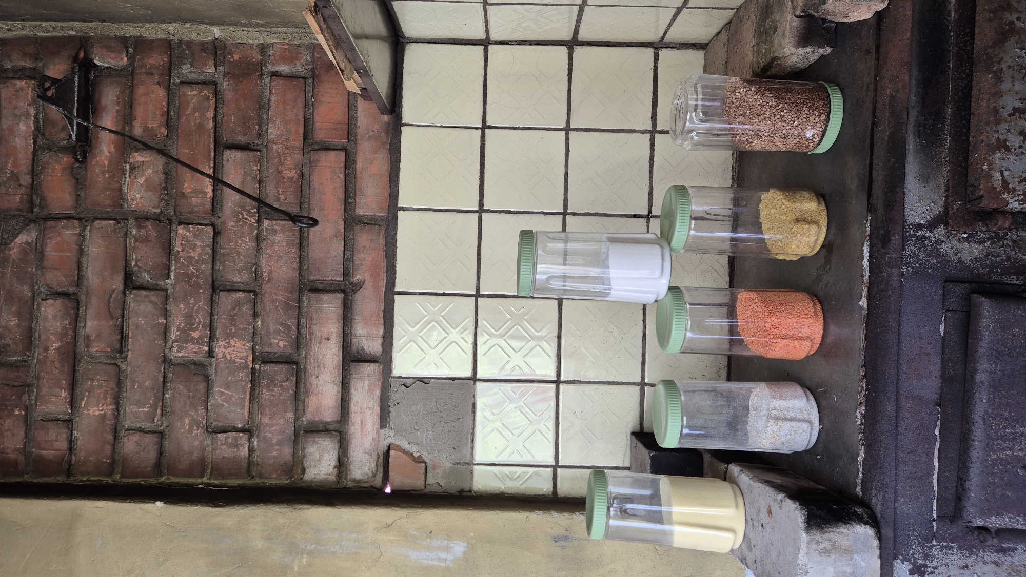 This is an image of a rustic kitchen area, likely in Mashezero, Russia. The scene features a brick wall on the left, some of which is covered in a light-colored tile with a diamond pattern. Several clear plastic jars with green lids are arranged on a flat surface on the right. These jars contain various dry goods: the top jar holds what appears to be buckwheat or a similar grain, the second contains yellow lentils or split peas, the third has orange lentils, and the fourth contains a white granular substance, possibly sugar or salt. The bottom two jars are partially visible and appear to contain more dry goods, one yellowish and the other white.
Next to the tiled area, on the right side of the image, is a dark metal structure that resembles an old stove or oven, with signs of rust and wear. A metal rod is attached to the brick wall, extending outwards. The overall impression is one of an older, functional kitchen, possibly in a rural or traditional setting. The lighting suggests it's daytime, but there are no strong indications of weather conditions. The image is oriented vertically. No people are visible. There is no visible text in the image.