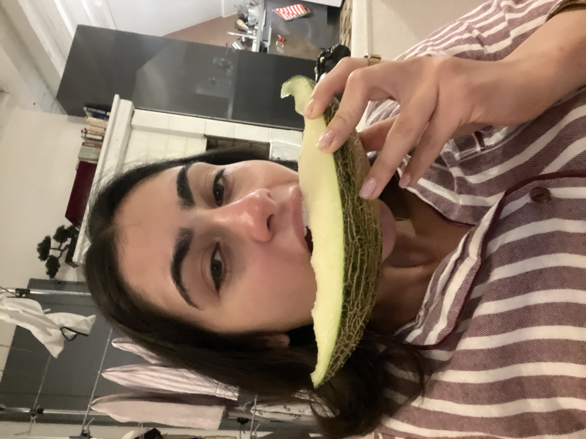 A woman is holding a slice of melon to her mouth, indoors. The woman has dark hair and is wearing a striped shirt. Background elements include a kitchen area with stainless steel appliances and a clothes rack. The location is indeterminate, and there's no verifiable indication that it is located in Paris, France.