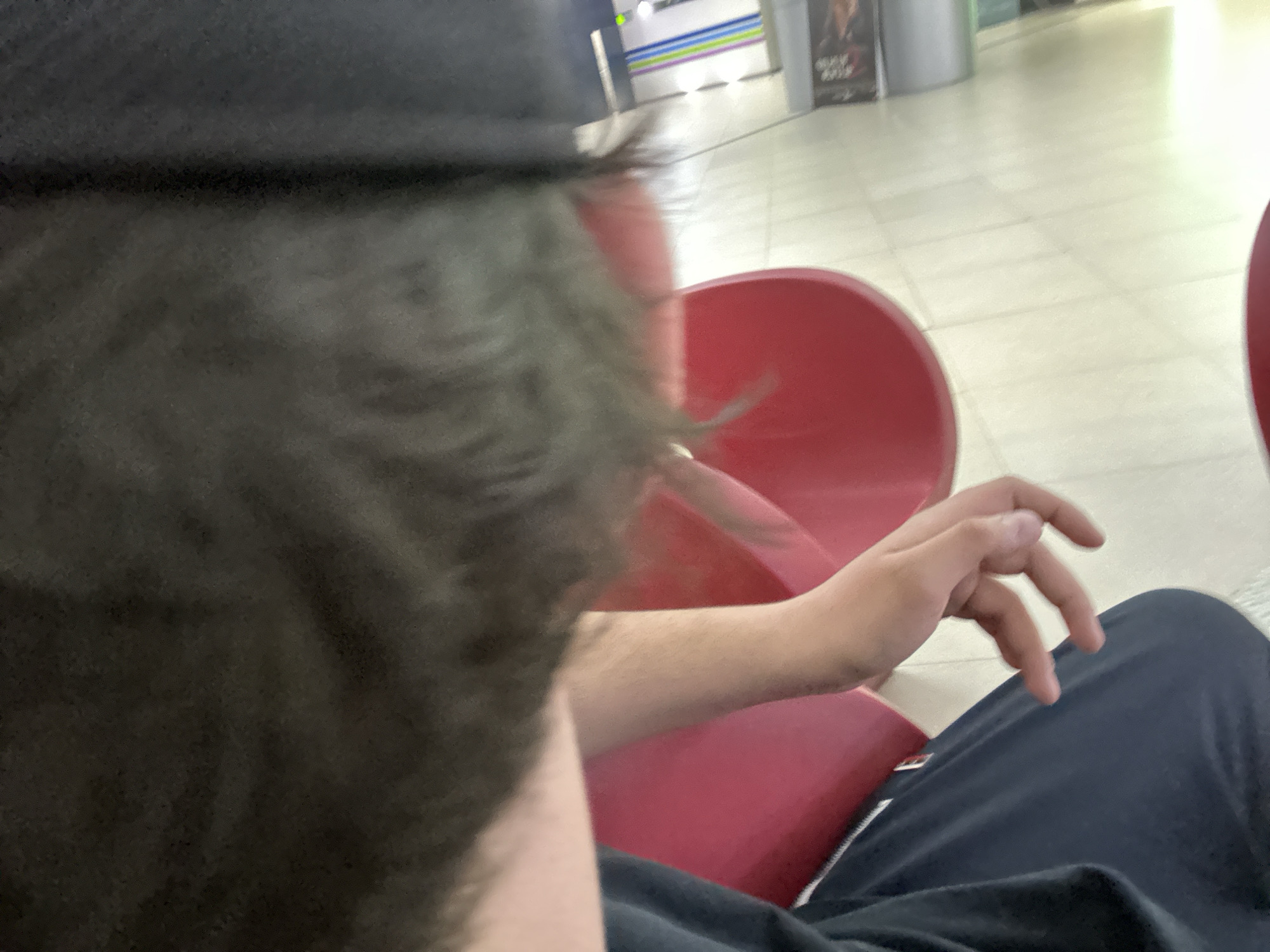 The image shows a person sitting in a red plastic chair, possibly at an airport or waiting area. The person is wearing a black cap and dark clothing. Part of their hand is visible. The location appears to be indoors with a tiled floor and a poster in the background. The city and exact location are unknown.
