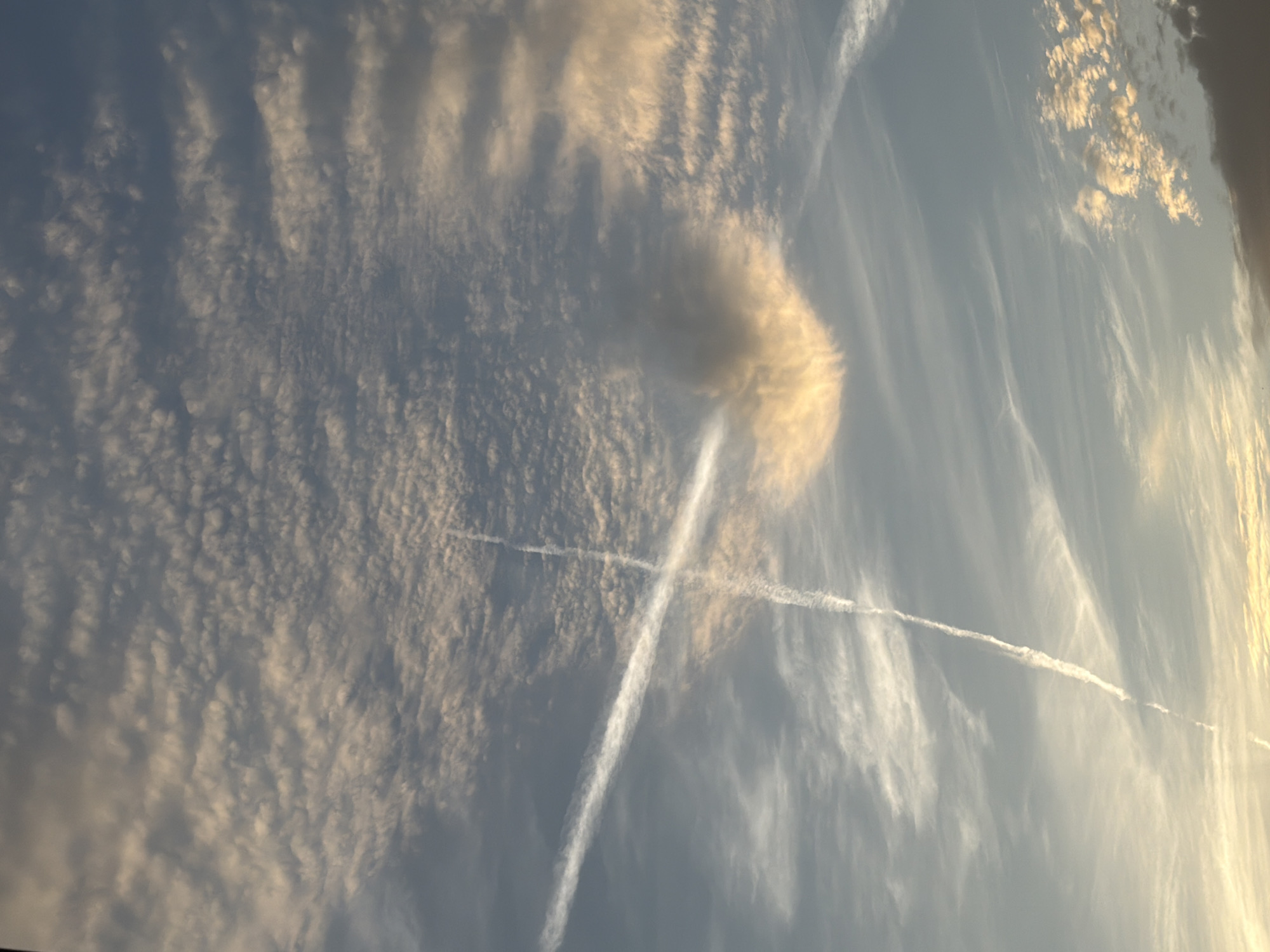 The image shows a sky with clouds and contrails. There are two prominent contrails that intersect, forming an "X" shape. The clouds are varied in type and density, with some appearing wispy and others more consolidated. The lighting suggests it might be near sunrise or sunset, with warm hues illuminating some of the clouds. The lower portion of the image shows a darker cloud bank, possibly indicating the horizon or a weather front. There are no people, subjects, or objects other than the atmospheric phenomena. The location in Budva, Montenegro, cannot be determined from the visual information in the image.