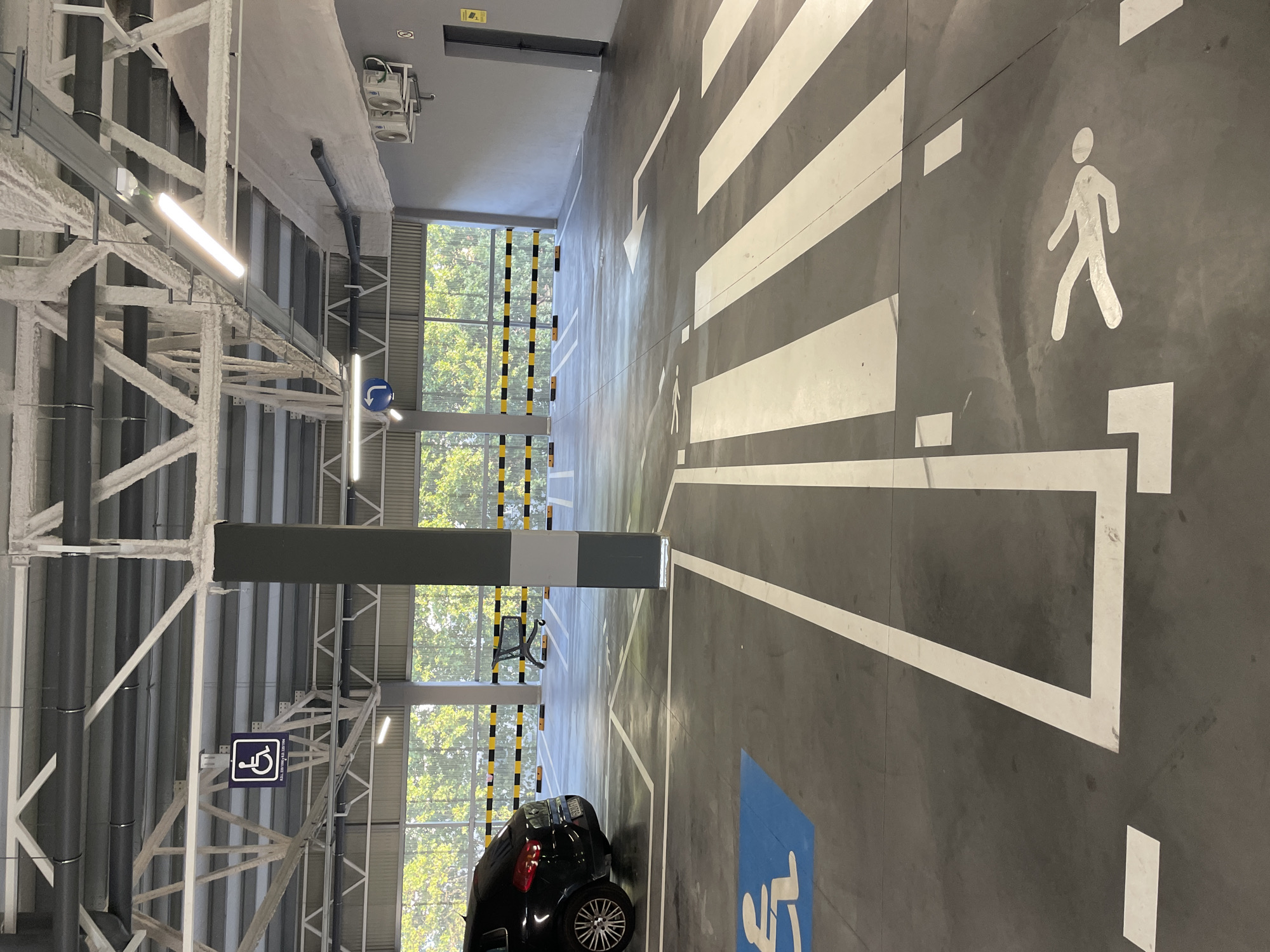 An overhead perspective shot shows a section of an indoor parking garage in La Línea de la Concepción, Spain. In the foreground, a pedestrian crossing with white stripes is visible on the gray concrete floor. To the left of the pedestrian crossing, a blue square with a white wheelchair symbol is painted on the ground, indicating a disabled parking spot. A portion of a black car is parked on the far left, its rear tire and fender visible. Further back, a yellow and black striped barrier runs across the width of the garage, separating the parking area from an open space. Beyond the barrier, trees and foliage are visible through an open-air section of the garage, suggesting it is a covered but not fully enclosed structure. Overhead, exposed ceiling structures with pipes, ventilation systems, and fluorescent lighting fixtures are present. A dark gray pillar stands prominently in the middle of the frame.