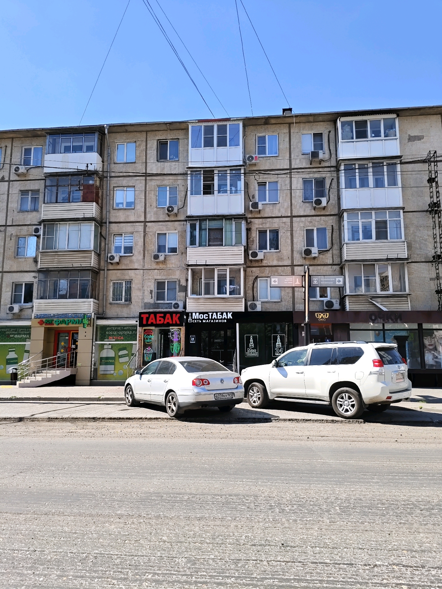 The image shows a street view in Rostov-on-Don, Russia, on a clear, sunny day. The primary subject is a multi-story apartment building with a weathered, concrete facade. The building has numerous windows and balconies, many of which are enclosed with glass. Air conditioning units are visible mounted outside many of the windows.
At the base of the building, there are several storefronts. From left to right, one store appears to be a pharmacy with prominent green signage that includes the text "ЮГ ФАРМ" (YUG FARM). Next to it is a tobacco shop with a red and black sign that reads "ТАБАК" (TABAK) and "МОСТАБАК" (MOSTABAK), featuring neon signage inside. Further to the right, there are other businesses with visible signs, including "ОККО" (OKKO) and "ОЧКИ" (OCHKI), suggesting an eyewear store.
Two white cars are parked on the street in front of the building. The car on the left is a sedan, and the car on the right is an SUV. The street in the foreground appears to be paved and shows signs of wear and tear. Power lines are strung across the top of the image, and a clear blue sky is visible above the building. There are no people clearly visible in the image.