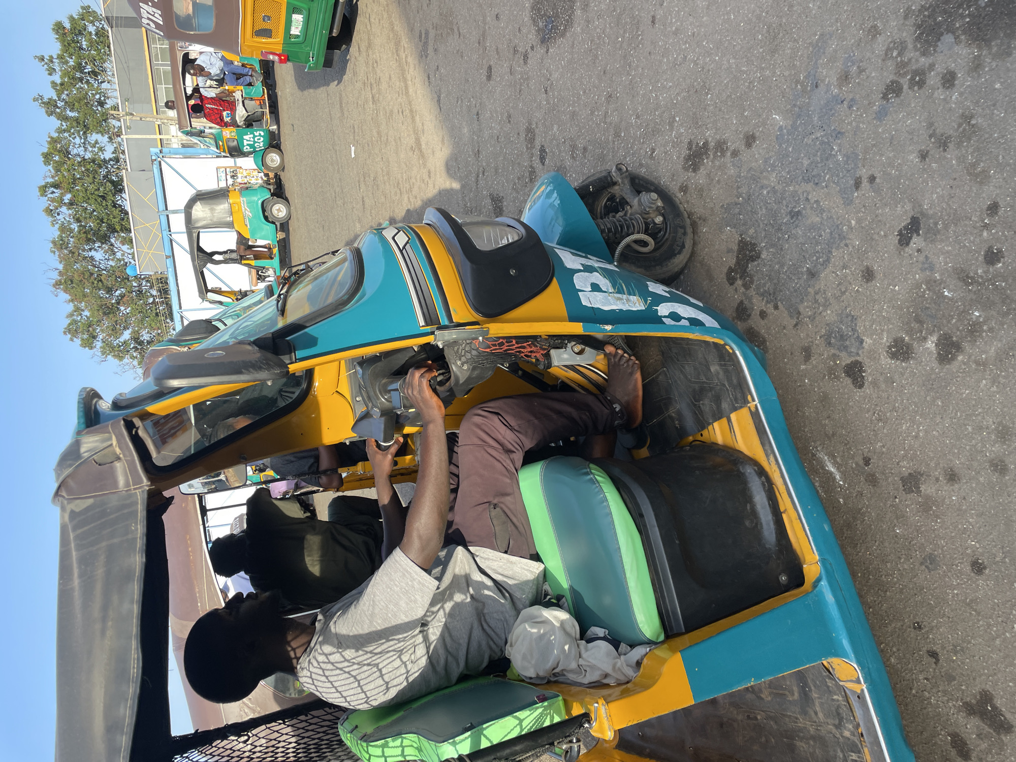 The image shows the interior and exterior of a three-wheeled motorized vehicle, possibly a rickshaw or auto-rickshaw, being driven or parked in a city.
**People:**
* At least one driver is visible at the steering mechanism, wearing brown pants and presumably sandals on his feet.
* Passengers are seated in the rear of the vehicle, at least one wearing a gray shirt.
* Other auto-rickshaws in the background appear to have drivers and/or passengers.
**Subjects and Objects:**
* The three-wheeled motorized vehicle is a prominent subject. It is painted yellow and blue.
* Other similar vehicles are parked nearby.
**Environment:**
* The location is likely a street or parking area within Jos, Nigeria, given the typical appearance of auto-rickshaws used for transportation in the city.
* The ground is paved and shows signs of wear and oil stains.
**Actions and Interactions:**
* The primary action is the operation (or preparation for operation) of the vehicle.