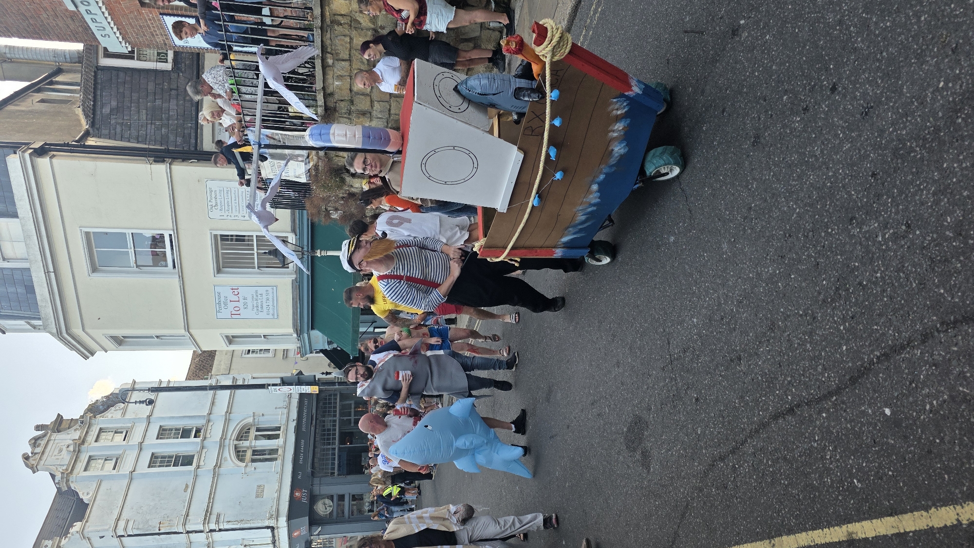 The image depicts a street scene in St Leonards, United Kingdom, during what appears to be a parade or festival. In the foreground, a man in a sailor costume with a false beard and pipe is pulling a small, boat-shaped float on wheels. The float is decorated with blue waves at the bottom and has a white cabin-like structure on top. A shark toy is attached to the side of the float. Behind him, other participants are visible, including someone in a shark costume and another in a grey shark costume. There are spectators lining the street on the right side of the image, some standing on a raised area. The buildings in the background are typical of a UK town, with shops and residential properties. The street is paved with asphalt. The overall atmosphere seems festive.