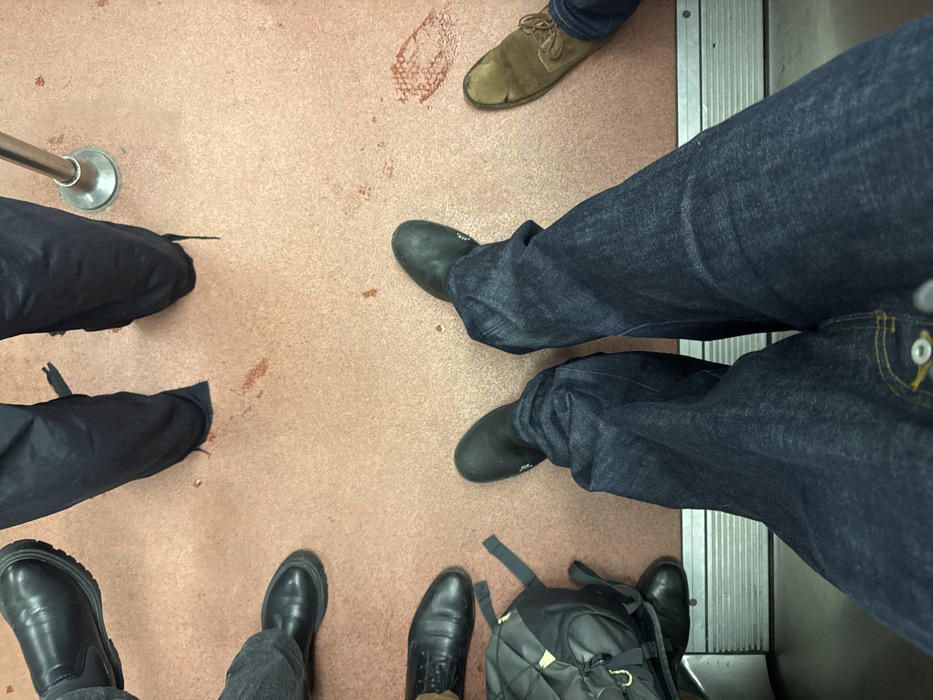 The image shows an overhead shot of a train or bus floor. Several people's feet and legs are visible, each wearing different shoes. One person wears suede shoes, another has black boots, and another has dark jeans. A backpack sits on the floor near someone's feet. The floor is a light brown color and has some smudges and debris on it. A silver rail can be seen on the left side. The edge of the open doors is visible on the right.