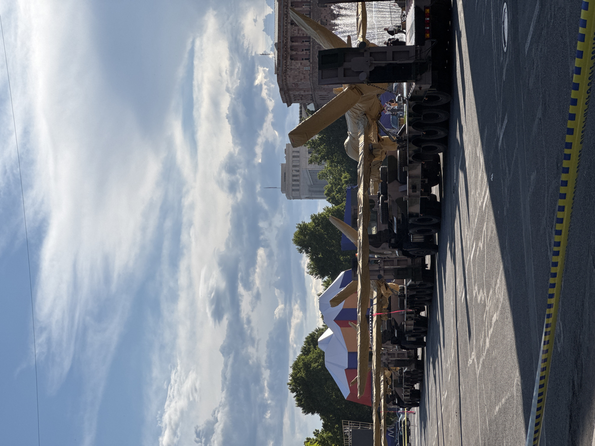 The image shows a row of military vehicles, likely missile launchers, parked on a street in Yerevan, Armenia. The vehicles are covered with protective tarpaulins. In the background are buildings, including the government building. A Russian flag is visible. The sky is partly cloudy. A yellow and blue striped barrier tape is visible in the foreground. The scene appears to be a military parade rehearsal.