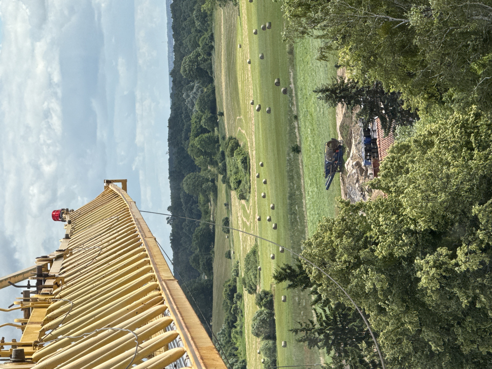 The image captures a scene in Mikołajki, Poland, likely a rural or semi-rural area. In the foreground, a large, yellow construction crane arm extends diagonally across the frame from the top left. A red warning light is visible on the crane. Below the crane, a lush green landscape unfolds, featuring rolling fields dotted with numerous round hay bales. A line of trees forms a dark green border in the background. Closer to the middle ground and slightly to the left, a dark-colored piece of construction equipment, possibly a crusher, sits on a patch of disturbed earth, surrounded by piles of rocks and soil. A red-tiled roof of a building is partially visible amidst the trees in the lower right quadrant. The sky is overcast with dramatic clouds. The overall impression is one of ongoing construction or agricultural activity within a natural setting.