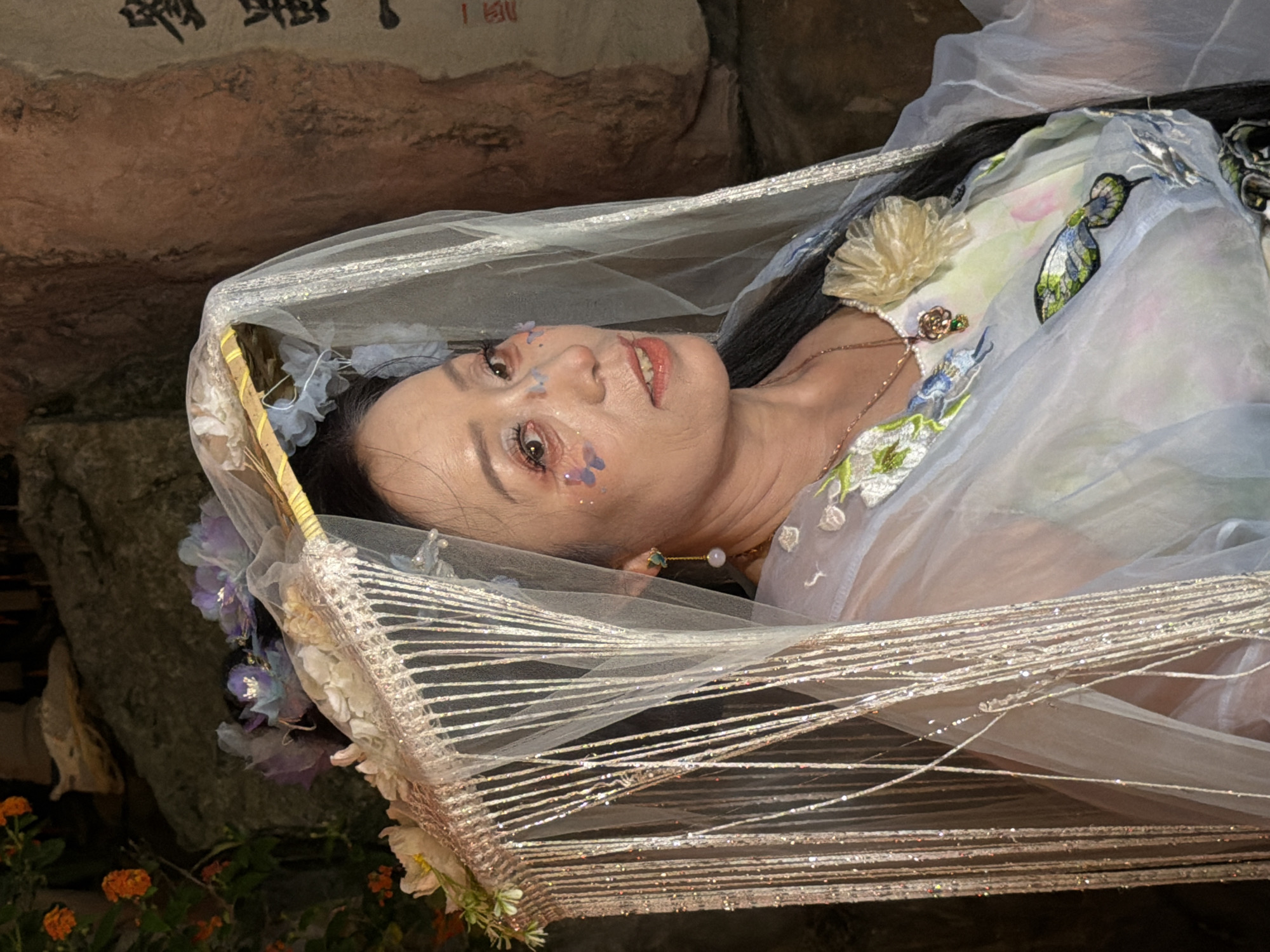 In Yuzhong District, China, a woman of Asian descent is depicted in a close-up portrait, adorned in elaborate attire. She wears a delicate veil, possibly part of a traditional headdress, embellished with floral accents and shimmering fringe. Her makeup includes subtle embellishments around her eyes and cheeks, resembling small butterflies. Her clothing appears to be a flowing, light-colored garment with embroidered floral and butterfly motifs. She is also wearing a necklace with a pendant. The background features a textured, rocky surface with some dark greenery and a portion of what appears to be a large Chinese calligraphy character. The overall impression is of a theatrical or ceremonial presentation.