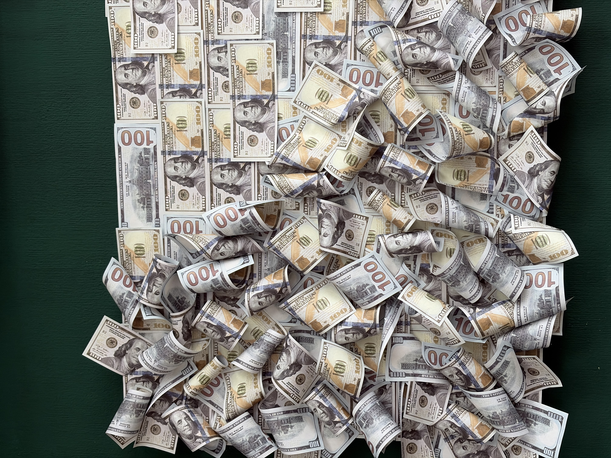 The image displays a dense pile of US 100-dollar bills, predominantly in a state of disarray, with many bills folded and some rolled. The bills are scattered across a dark green textured surface, possibly a table or fabric, which occupies the bottom portion of the frame. The upper part of the image shows a plain dark green background. The lighting appears to be from above, casting subtle shadows. No people, specific locations in Chaikovsky, Russia, or any actions or interactions are depicted. The scene is solely focused on the arrangement of the currency.
