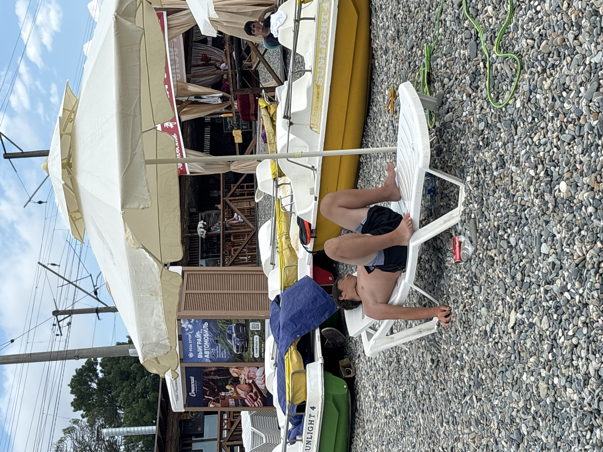 A young man with dark hair and dark swim trunks is lounging on a white plastic beach chair on a pebble beach. He is lying on his back with his head tilted to the side and his eyes closed, appearing to be asleep or resting. His legs are bent at the knees and his feet are propped up. A red and white soda can is lying on the ground near his feet. In the background, several pedal boats are lined up on the pebble beach, some covered with blue tarps. A large white umbrella shades a seating area. Beyond the boats and umbrella, there is a building with wooden structures and awnings, with signs and posters visible. Power lines and utility poles are visible against a cloudy sky. The scene appears to be a beach resort or recreational area in Sochi, Russia, possibly along the Black Sea coast.