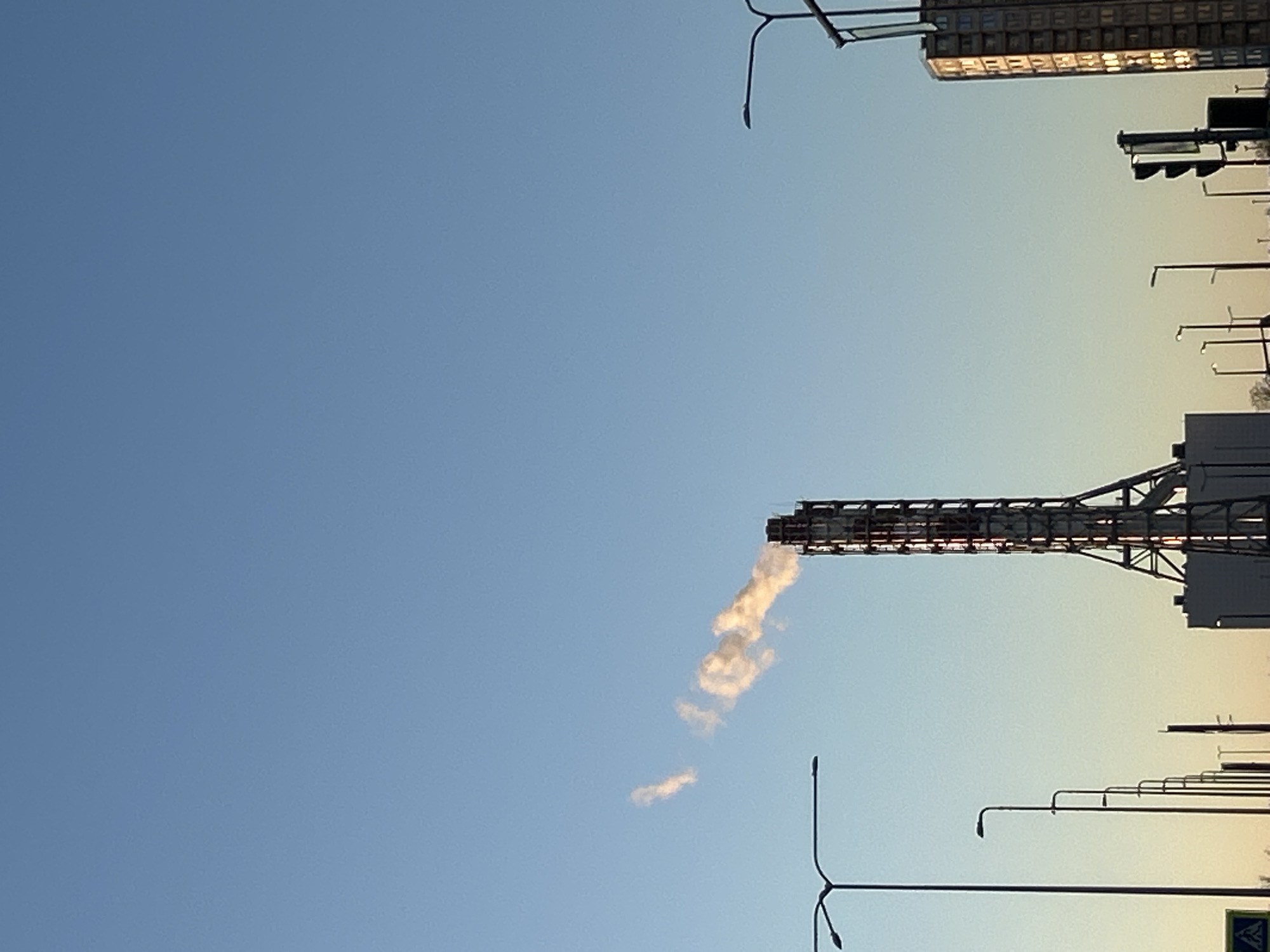 This image captures an urban or industrial landscape in Saint Petersburg, Russia, under a clear sky. The scene appears to be set during late afternoon or early morning, as indicated by the gradient in the sky, transitioning from a deeper blue overhead to a lighter, slightly yellowish hue towards the horizon.
The most prominent subject is a tall, dark, metal-framed industrial structure, resembling a chimney or flare stack, positioned towards the right side of the frame. From its top, a plume of light-colored smoke or vapor is being emitted, extending diagonally upwards and to the left, gradually dissipating into thinner wisps against the blue sky.
In the upper right background, a modern-looking building with numerous illuminated or highly reflective windows is visible, suggesting contemporary architecture. Below it, a traffic light structure can be seen. Multiple dark, silhouetted streetlights or lamp posts with distinctive curved arms line what appears to be a road or street, stretching from the middle-right to the bottom-right of the image. A small portion of a blue and white pedestrian crossing sign is visible in the bottom right corner, confirming an urban street setting. No people are visible, and no discernible text appears in the image. The weather is clear, with no natural clouds, only the industrial emission.