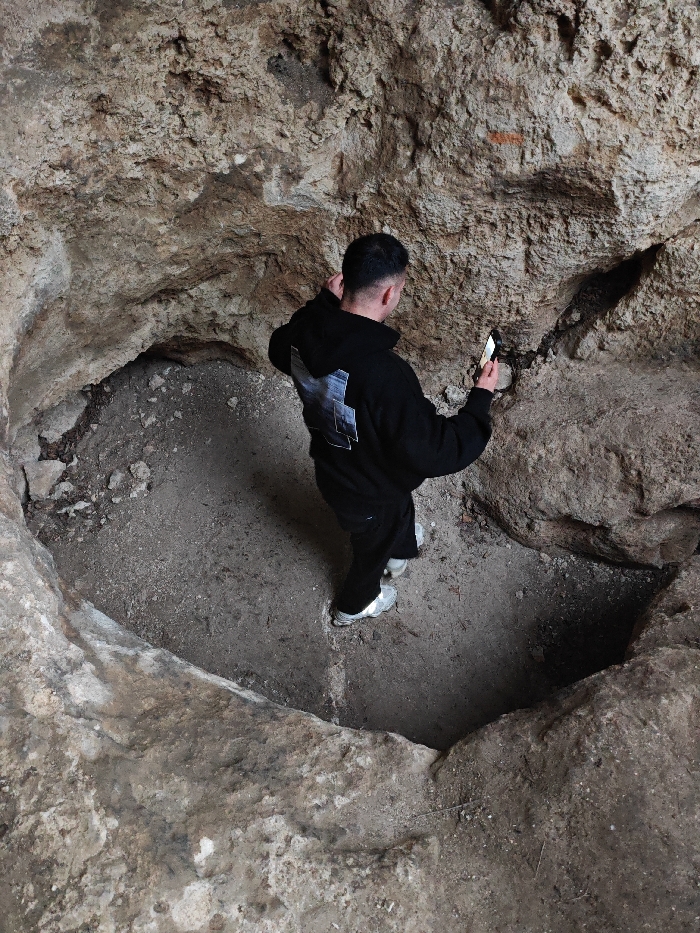 An overhead view captures a single male individual standing within a rough, excavated, rock-lined pit in Shumen, Bulgaria. The subject is seen from behind, wearing a black hooded sweatshirt with a light-colored, multi-paneled graphic on the back, dark trousers, and light-grey athletic shoes. He holds a white smartphone in his right hand, pointed towards the textured, stratified rock wall directly ahead of him, with the phone's screen emitting light onto the surface. The pit's irregular walls consist of light brown and grey rock, exhibiting various crevices and uneven surfaces. A distinct, dark, vertical fissure is visible on the rock wall to the subject's right. The ground within the pit is unpaved, composed of packed earth and scattered debris, bisected by a faint, lighter diagonal line.