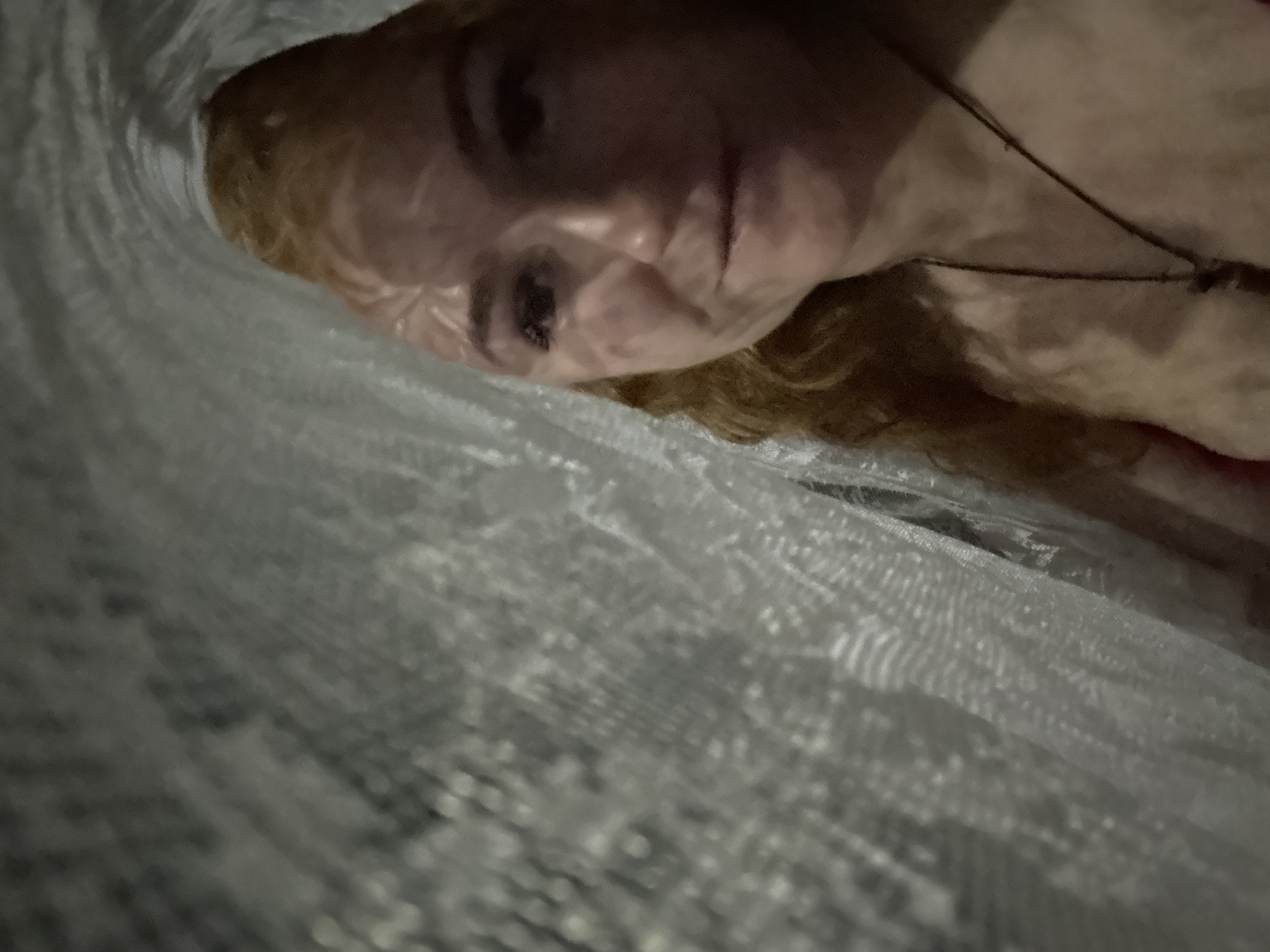 A person with red hair and fair skin is partially visible through a light gray, semi-transparent fabric or net. The person's face is the clearest part of the image, showing a portion of the forehead, an eye, a nose, and part of the mouth. A necklace is visible on the person's chest. The background is indistinct and dark. The location of the city of Chaikovsky, Russia cannot be verified from this image.