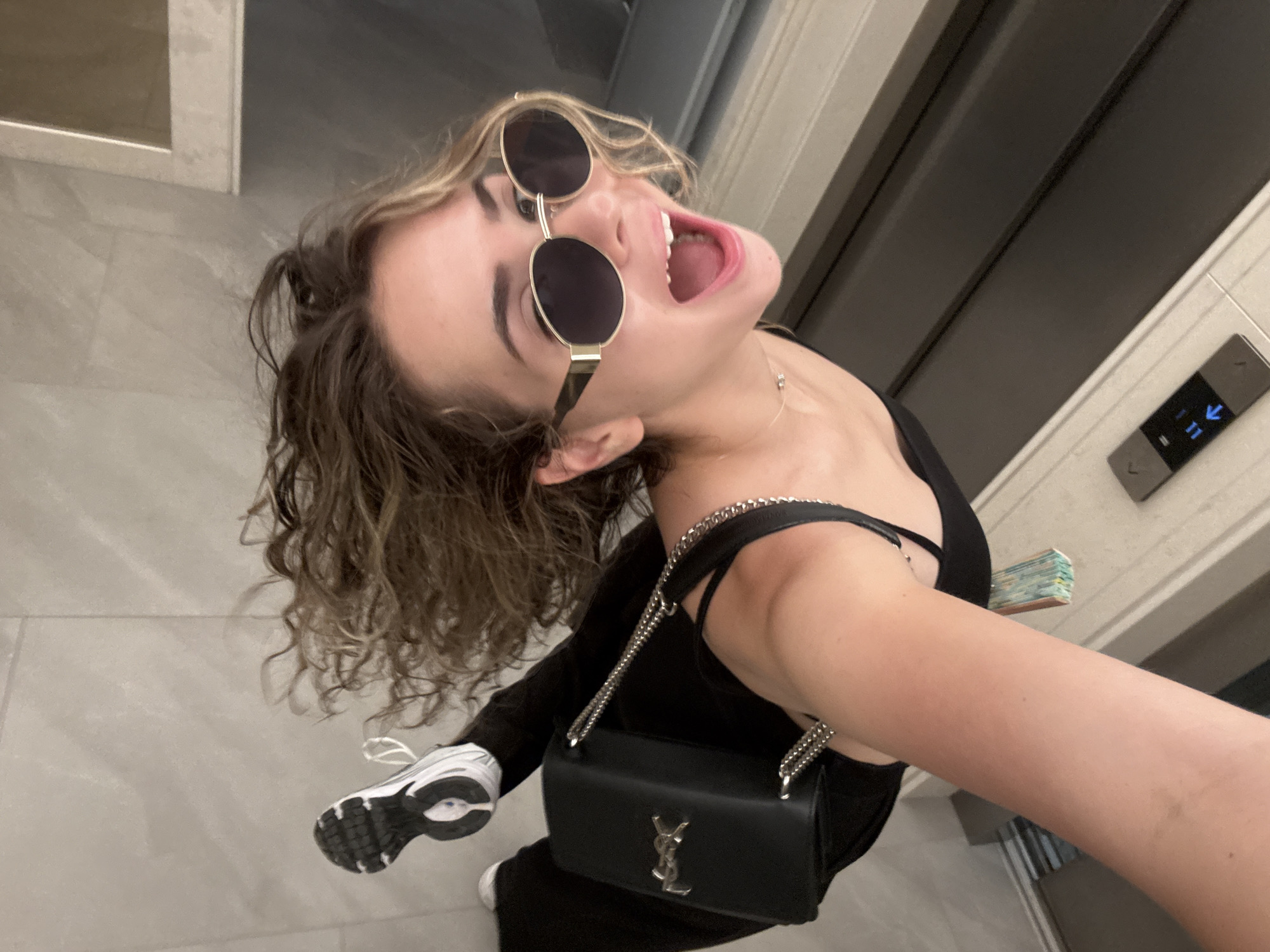 An upward-angled, low-perspective selfie captures a light-skinned woman indoors. She has curly, light brown hair, wears round, dark-lensed sunglasses with gold frames positioned on her nose, and has her mouth open wide, as if mid-laughter or shouting. A second pair of sunglasses rests on her head. She is adorned with a thin silver necklace featuring a small pendant and is dressed in a black top and black pants. A black YSL-branded shoulder bag with a silver chain strap is slung across her body. Her left foot, wearing a white athletic shoe with a black sole, is partially visible. In her right hand, she holds a fanned stack of green and blue banknotes. The setting features a light gray tiled floor with thin grout lines. Behind her are dark gray vertical panels, likely an elevator shaft or wall, and to the right, an elevator control panel on a light wall displays directional arrows and the number "11," indicating the eleventh floor. The scene is located in Sofia, Bulgaria.