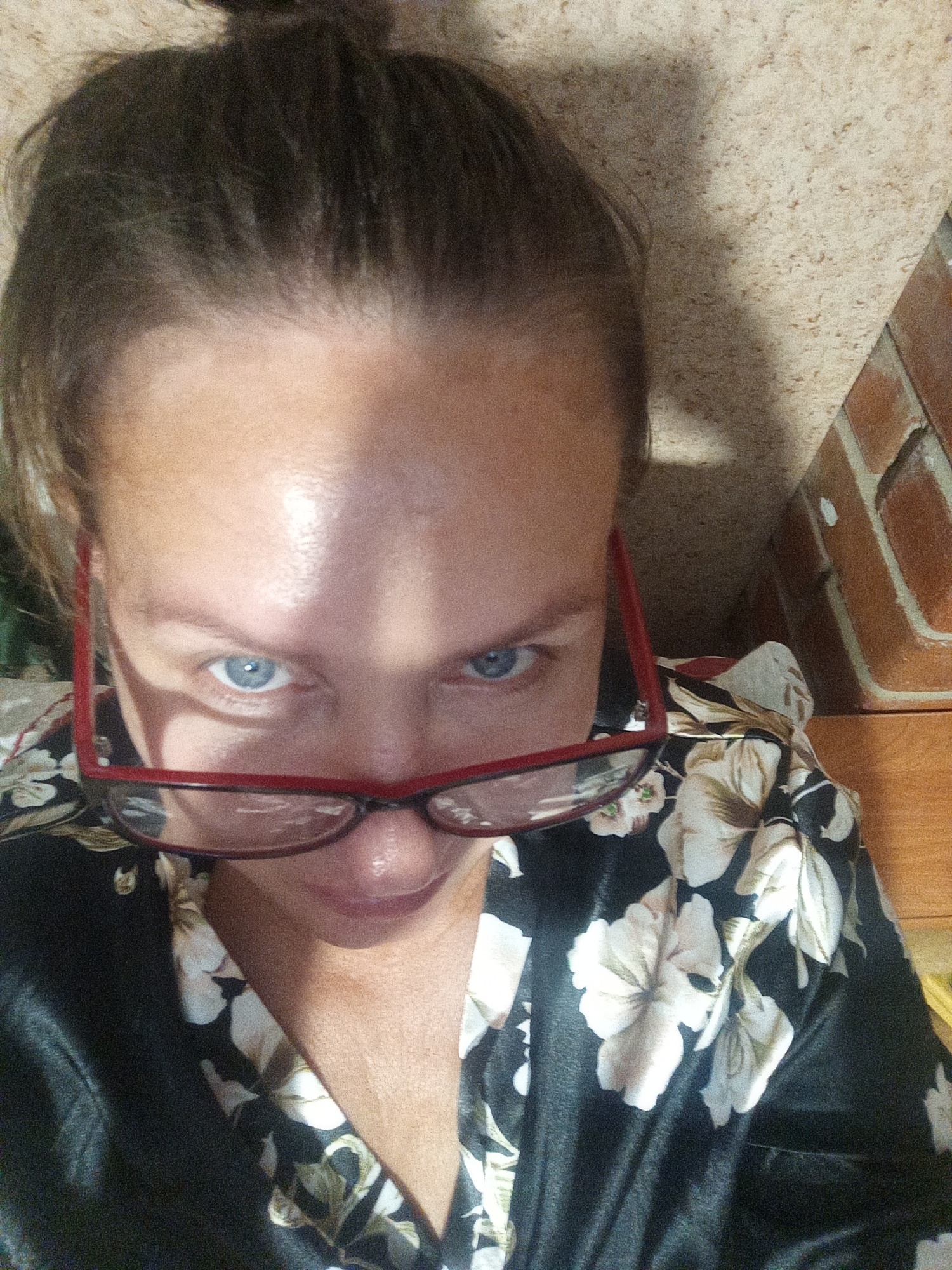 A close-up, slightly angled, indoor selfie of a woman with light skin and brown hair pulled up in a bun is shown. She is wearing red-framed glasses and a black robe with a white floral pattern. Her blue eyes are looking directly at the camera, and her mouth is slightly open. The background consists of a textured wall and brickwork, suggesting an interior environment, possibly in Novopokrovka, Russia.