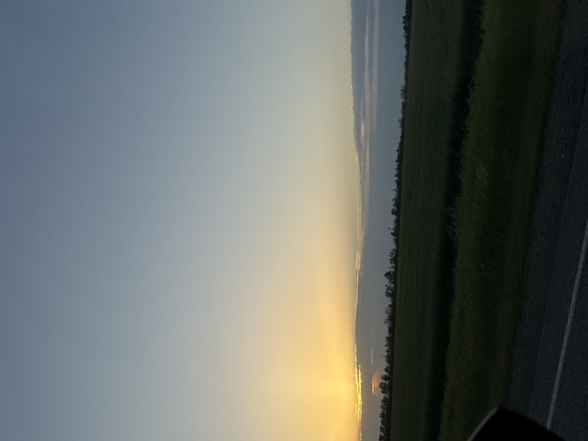 The image depicts a landscape at sunset. On the right side, there is a dark green field bordered by trees. To the right of the field, a paved road is visible with a white line. The left side of the image shows the sky transitioning from blue to yellow near the horizon, where the sun is setting. The location is Unknown, Unknown.