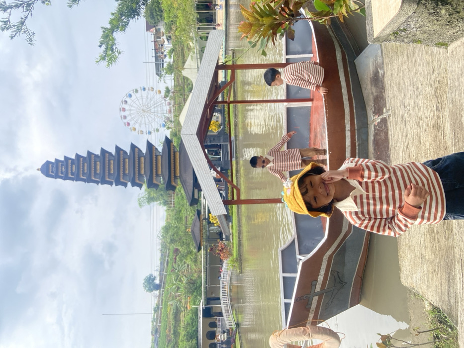 A whimsical scene captures a young girl in a striped shirt and yellow hat, positioned near a boat-like structure. Two other children, also in striped shirts, are seen on the boat. In the background, a tall, multi-tiered building and a Ferris wheel add to the charm. The setting appears to be a recreational area with a pond or water feature reflecting the sky. The image is slightly tilted, enhancing the playful atmosphere.