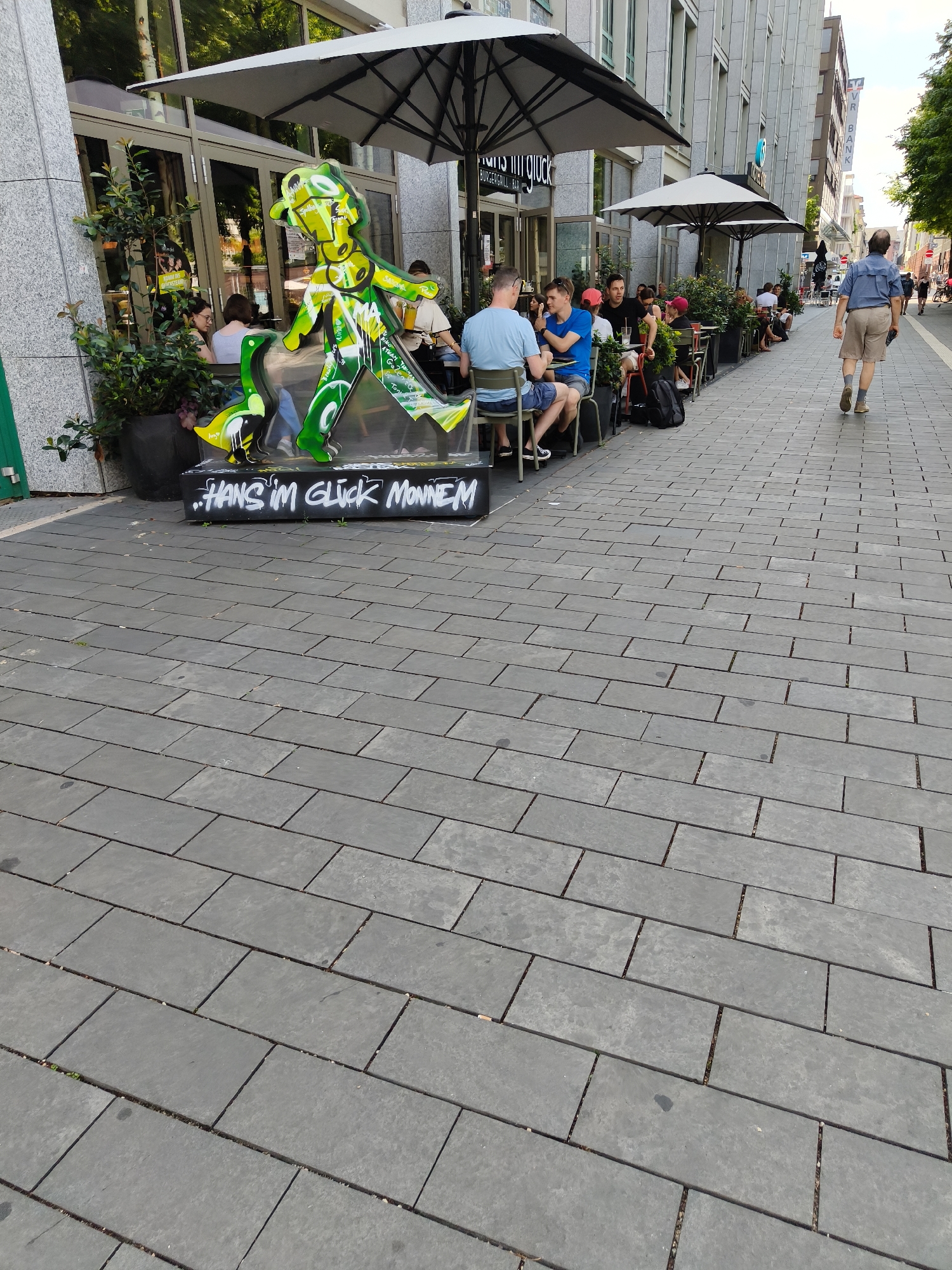 The image depicts a street scene in Mannheim, Germany, likely in an area with outdoor dining. In the foreground, the paved sidewalk is composed of dark gray rectangular stones. A large, green, abstract sculpture of a figure with outstretched arms and legs stands on a black base with white graffiti that reads "HANS IM GLÜCK MONNEM". Behind the sculpture, a restaurant with large glass windows and awnings is visible. Several people are seated at tables outside the restaurant, enjoying drinks and meals under large umbrellas. Some individuals are walking along the sidewalk in the background, including a man in a blue shirt and beige shorts walking away from the camera. The architecture of the surrounding buildings suggests an urban setting. The overall impression is a lively, commercial street with people engaged in leisurely activities.