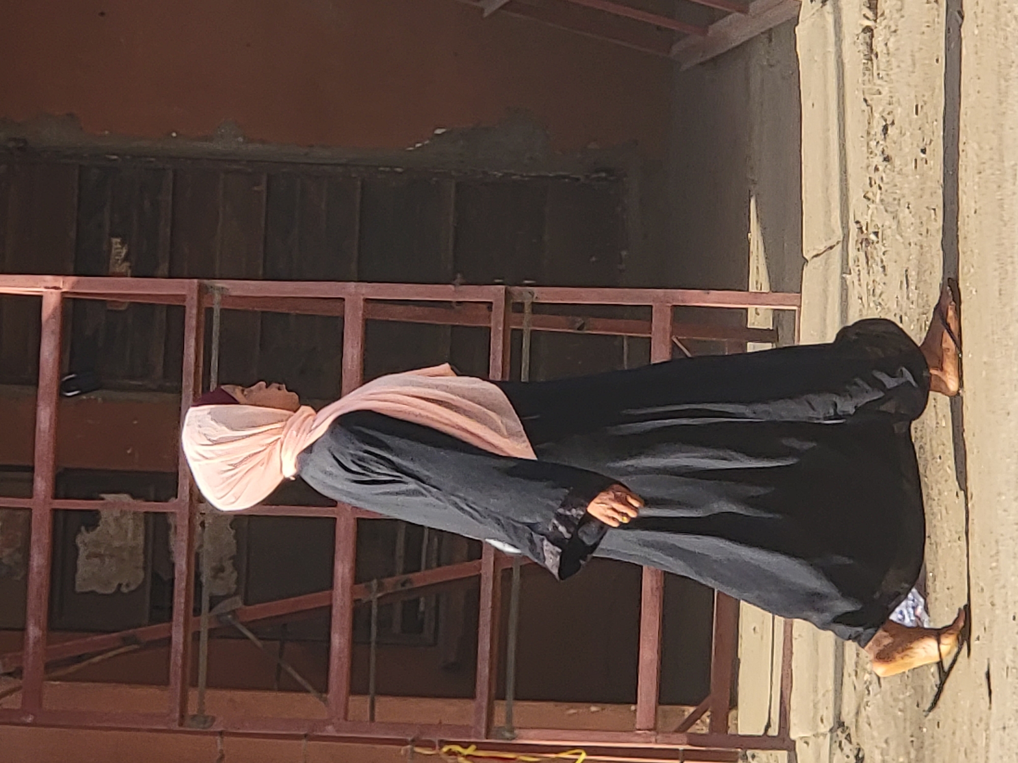 The image captures a full-body view of a person standing outdoors on a bright, sunny day in what appears to be a developing or construction area in Maiburiji, Nigeria.
The subject is a person, likely female given the attire, standing upright and looking slightly upwards and to their left. They are dressed in a long, dark garment, possibly an abaya, with long sleeves that cover their arms. A light pink or peach-colored head covering, resembling a hijab, is worn, concealing their hair. Their feet are visible, wearing simple dark sandals, and their skin tone appears medium to dark brown.
Behind the person is a reddish-brown metal framework or scaffolding structure, which occupies a significant portion of the mid-ground. Beyond this framework, a darker, shadowed interior space or wall is visible, suggesting an unfinished building or structure. To the right of the person, a rough-textured, light-colored concrete or stucco wall stands, illuminated by direct sunlight, casting subtle shadows. At the bottom edge of the image, yellow electrical wires or a rope are visible on the ground near the metal structure. The bright sunlight indicates it is daytime, and the overall scene suggests a warm climate. No discernible text is visible in the image.