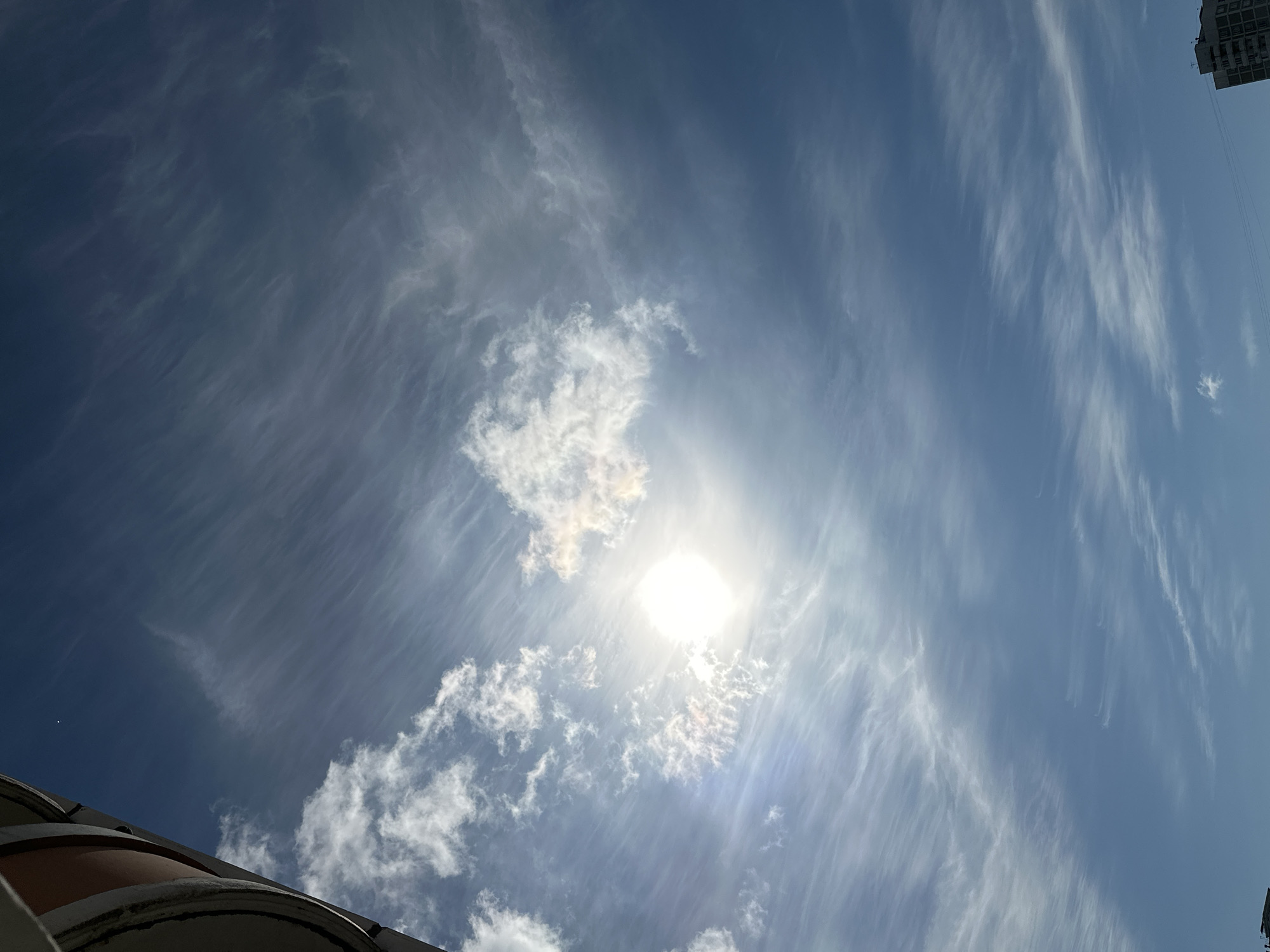 A clear blue sky with wispy, cirrus clouds stretches across the frame. The sun is visible, partially obscured by a thin cloud formation, casting a bright glare. The edge of a building with a rounded, curved section and alternating light and dark bands is visible in the upper left corner, suggesting an urban environment. In the lower right corner, a portion of a multi-story apartment building is visible against the sky. The scene appears to be an outdoor view from a city, likely Podolsk, Russia, based on the context provided.