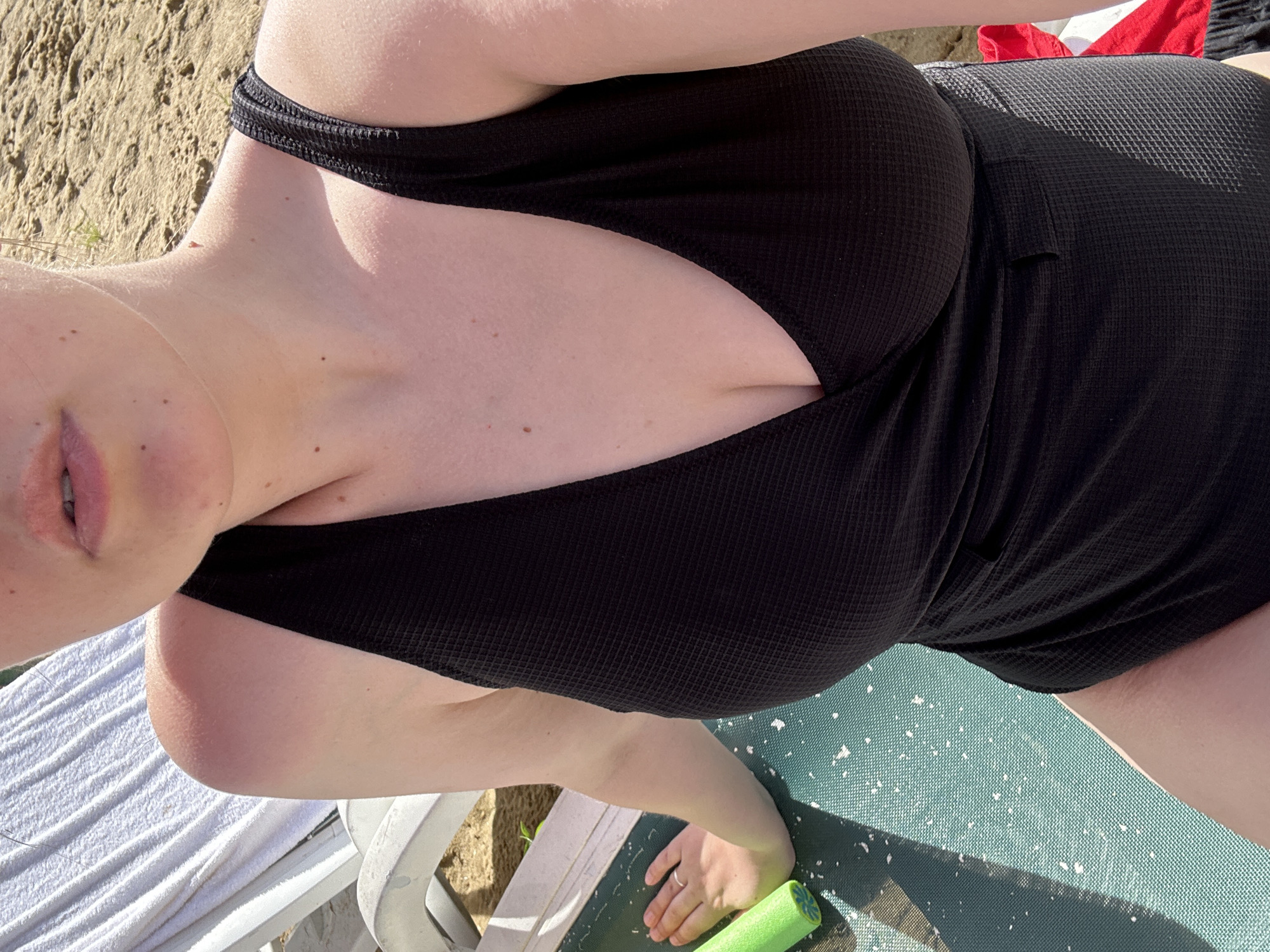The image shows a close-up of a woman wearing a black one-piece swimsuit. She is sitting on a green lounge chair with a white towel draped behind her. A sandy area is visible in the background. The woman's upper body, including her shoulders, chest, and part of her face, is in focus. Her skin appears sun-kissed, with some areas of redness on her shoulder. Her lips are slightly parted, and she is wearing a ring on her left hand. Two green pool noodles are partially visible on the lounge chair next to her hand. The lighting suggests it is a sunny day. The specific location within the city of Юбилейный, Russia, cannot be determined from the visual information provided.