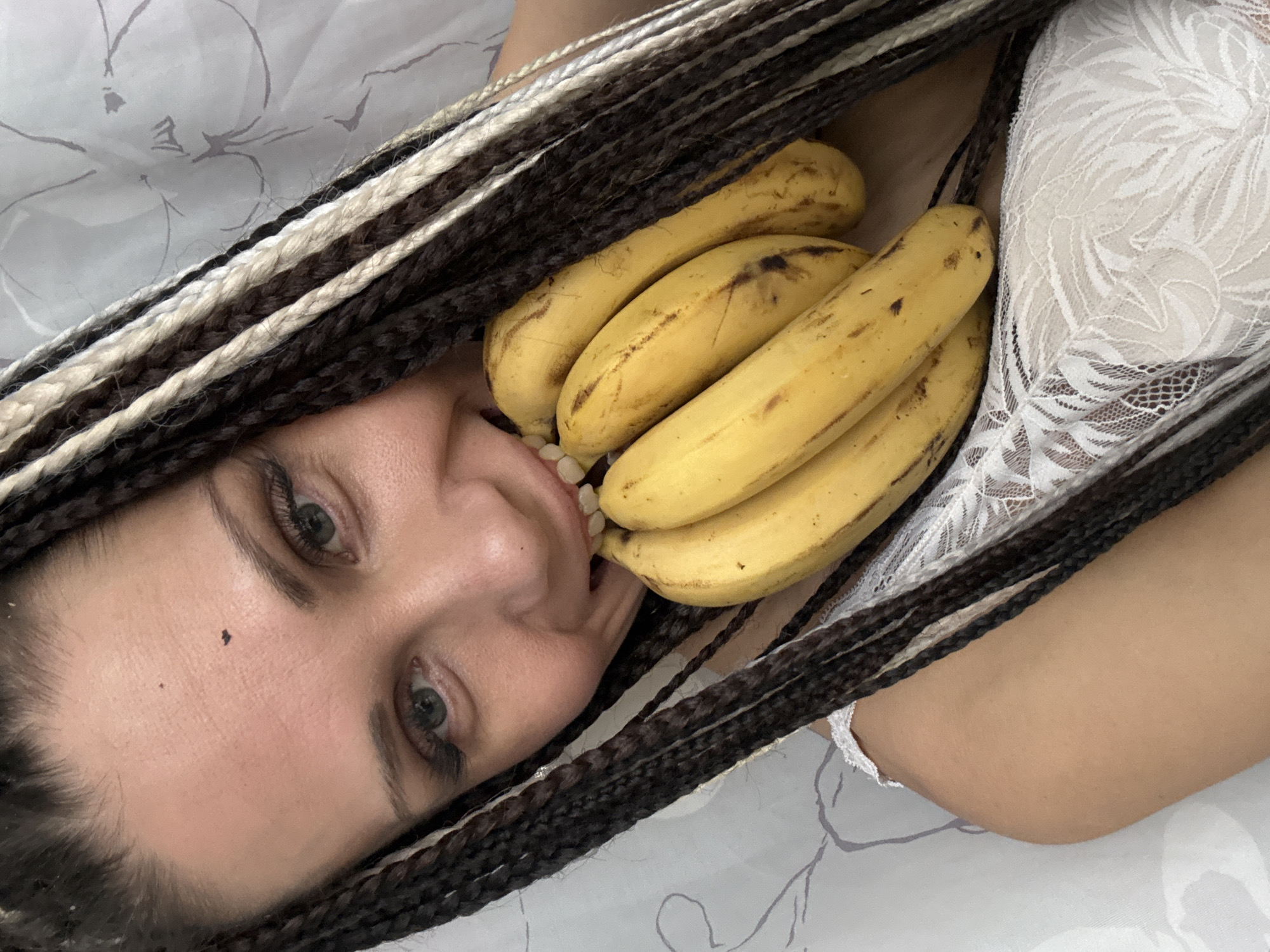 A woman lies supine on a bed covered with a patterned white sheet. Her face is close to the camera, revealing light-colored eyes, brown eyebrows, and dark braided hair with blonde strands. She is holding a bunch of ripe bananas in front of her, with one or two bananas partially in her mouth. She is wearing a white lace top. The location of the image is unknown and there is no evidence that this image was taken in Rusanovo, Russia.