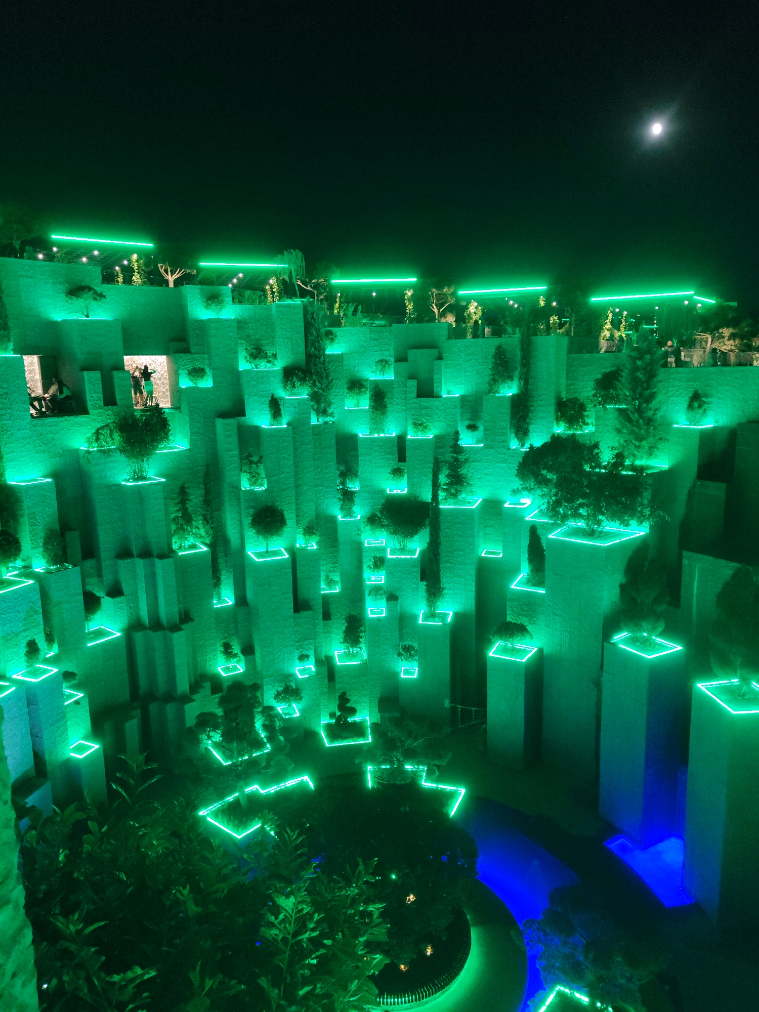 The image is an aerial, nighttime view of an outdoor installation in Krasnodar, Russia. The scene is dominated by illuminated, tiered structures resembling terraced gardens or a stylized rock formation. Each tier and planting area is outlined with bright green neon lights, creating a vibrant, artificial glow against the dark sky. Small trees and plants are strategically placed on various levels within these illuminated squares. In the upper right portion of the sky, a full moon is visible. In the foreground, lush green foliage, also outlined with green neon, fills the bottom of the frame. A dark, reflective body of water, illuminated by blue lights, winds through the lower part of the installation. Several indistinct figures, appearing as silhouettes, are visible on some of the upper platforms, suggesting human presence and interaction within the illuminated landscape. The overall impression is one of a modern, artistic, and possibly entertainment-oriented space designed for nighttime viewing.