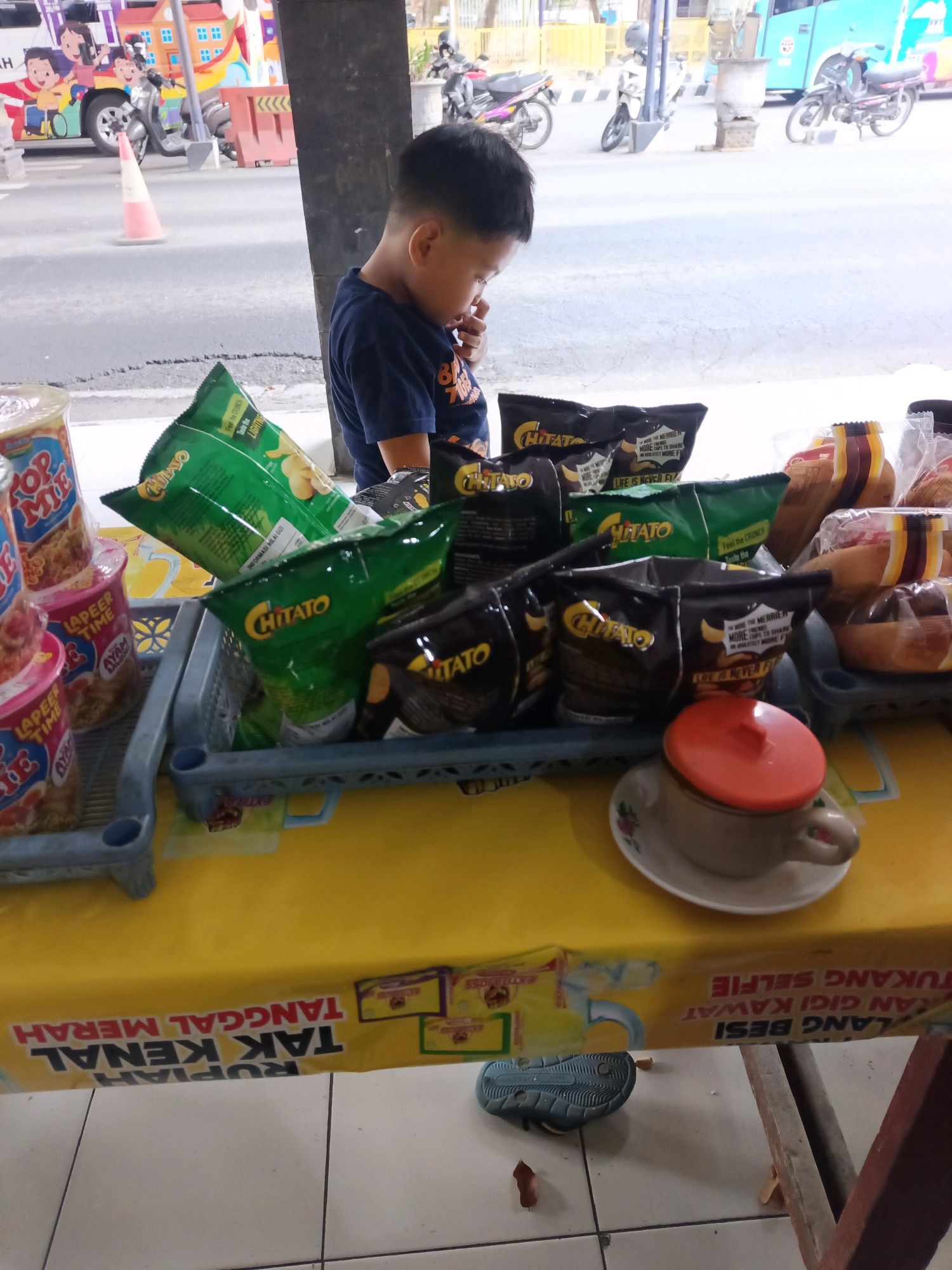 A young boy with short black hair, wearing a dark blue t-shirt with orange lettering, stands to the right of the frame, looking down. He is positioned behind a table covered with a bright yellow cloth. On the table, a variety of snacks are displayed, including several bags of "Chitato" potato chips, some in green and others in dark brown packaging. There are also stacks of instant noodle cups labeled "Top Mie" and "Lapeer Time." A small white teacup with a red lid sits on a saucer to the right of the snacks. In the background, a street scene is visible with traffic, including motorcycles and a bus. The street is paved, and there are buildings and a traffic cone visible. The overall impression is that of a small roadside stall. The text on the yellow tablecloth suggests the location is in Kediri City, Indonesia, with phrases like "RUPIAH TAK KENAL TANGGAL MERAH" (Rupiah Knows No Red Dates) and "TUKANG SELFIE" (Selfie Seller), indicating a commercial setting.